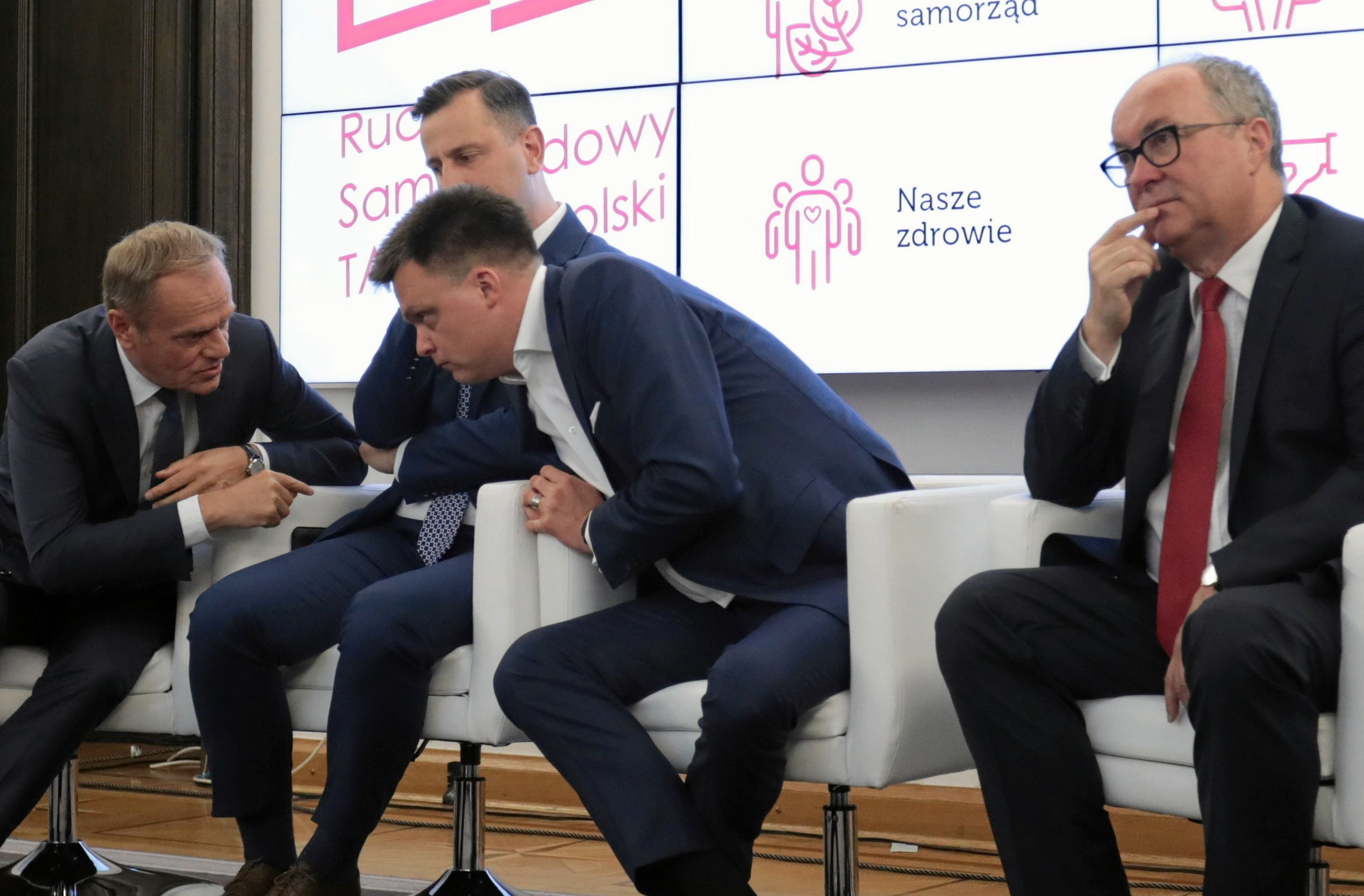 Czterej męzczyźni Tusk, Kosiniak-Kamysz, Hołownia i Czarzasty siedzą w fotelach. Tusk i Hołownia rozmawiają pochylając sie do siebie nad kolanami Kosiniaka-Kamysza