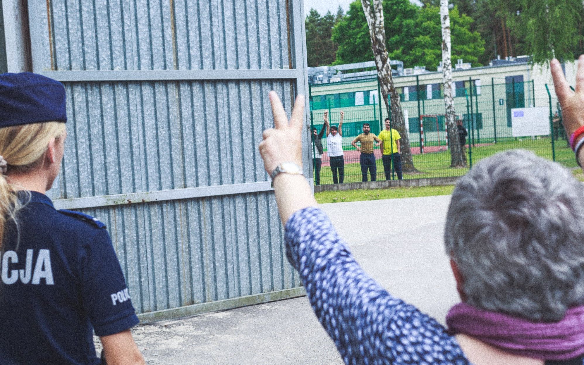 Ludzie stoją przed wysoka bramą, rece podnoszą w znaku zwycięstwa V