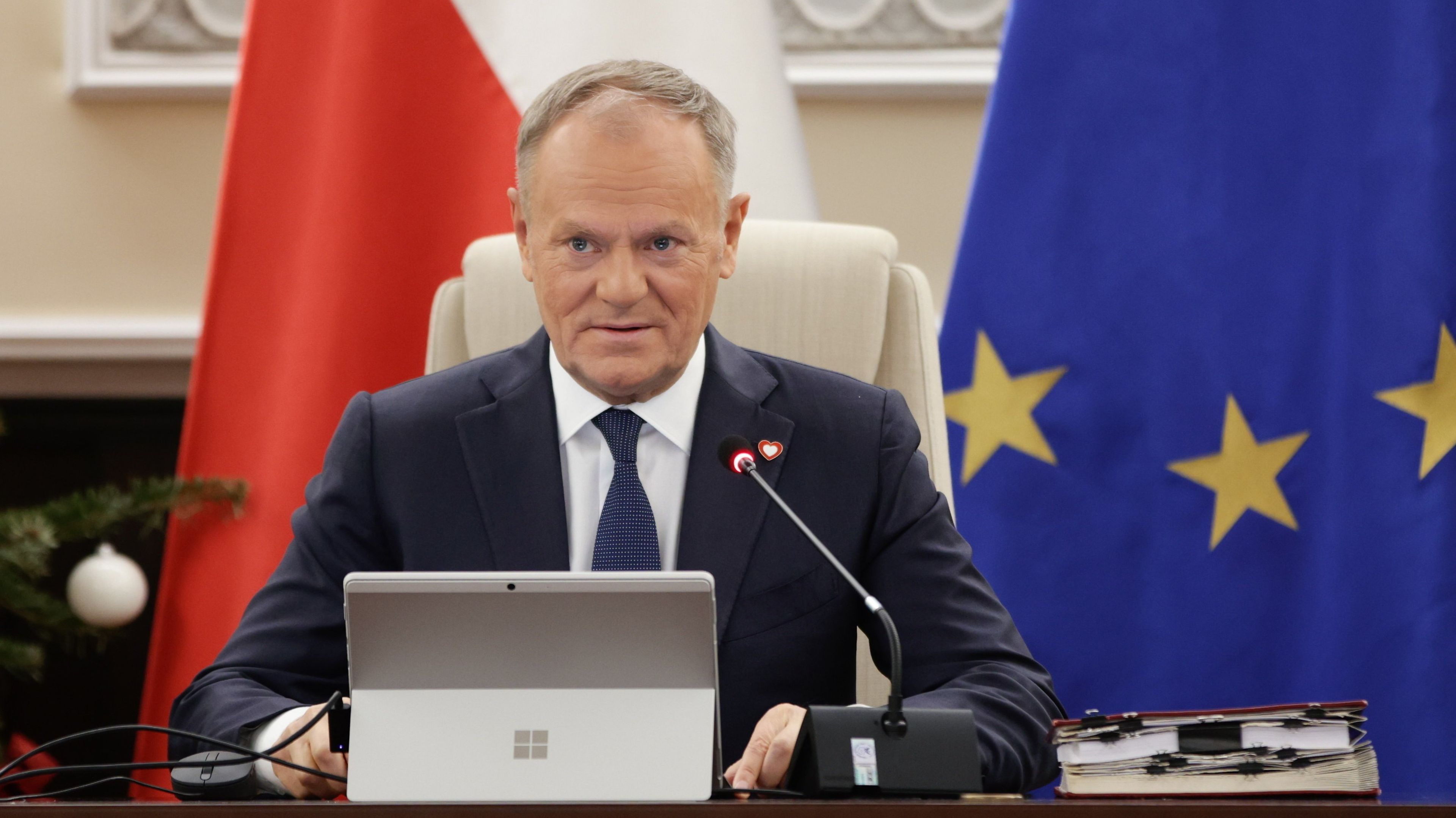 Mężczyzna, Donald Tusk, siedzi i premawia, za nim flagi biało-czerwona i unijna z gwiazdkami