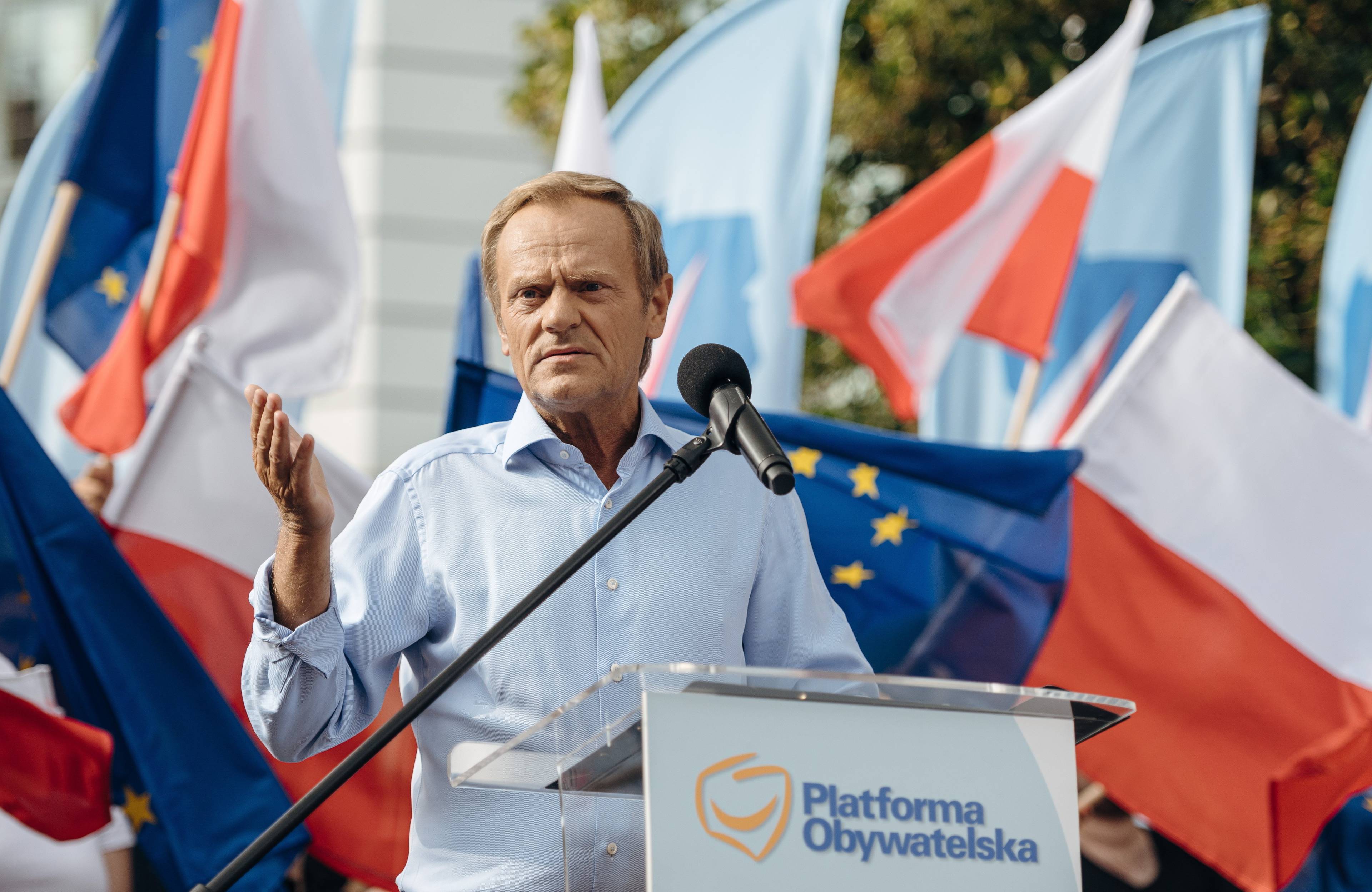 Donald Tusk w jasnej koszuli stoi za mównicą podczas demonstracji. Za nim powiewają flagi Polski i Unii Europejskiej