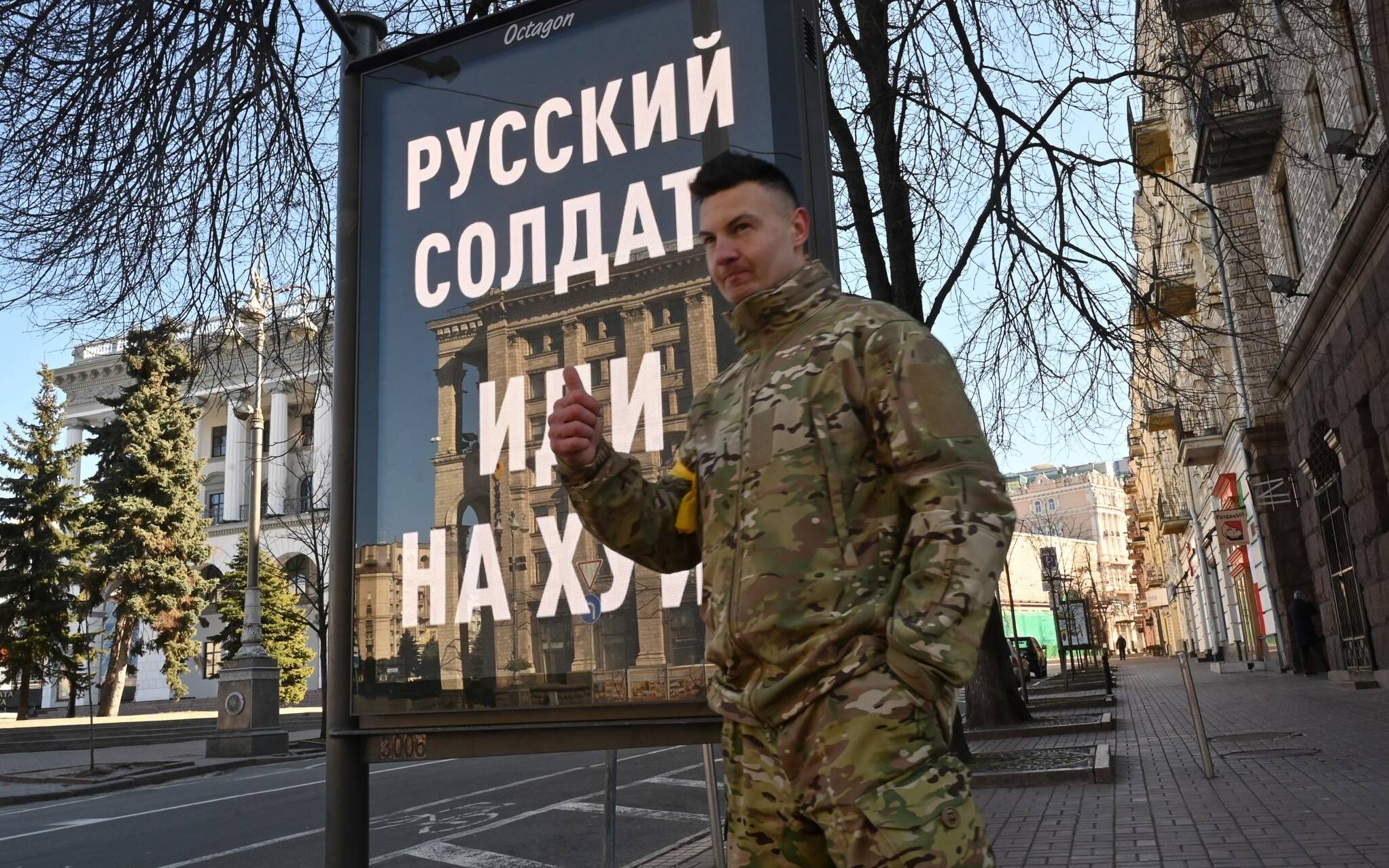 A fighter of the Ukrainian Territorial Defence Forces, the military reserve of the Ukrainian Armed Forces, poses for a picture by an advertising board with a message reading "A Russian soldier, go fuck yourself" at Kyiv's Independence Square on February 28, 2022. - The Russian army said on February 28, 2022, that Ukrainian civilians could "freely" leave the country's capital Kyiv and claimed its airforce dominated Ukraine's skies as its invasion entered a fifth day. (Photo by Sergei SUPINSKY / AFP)
