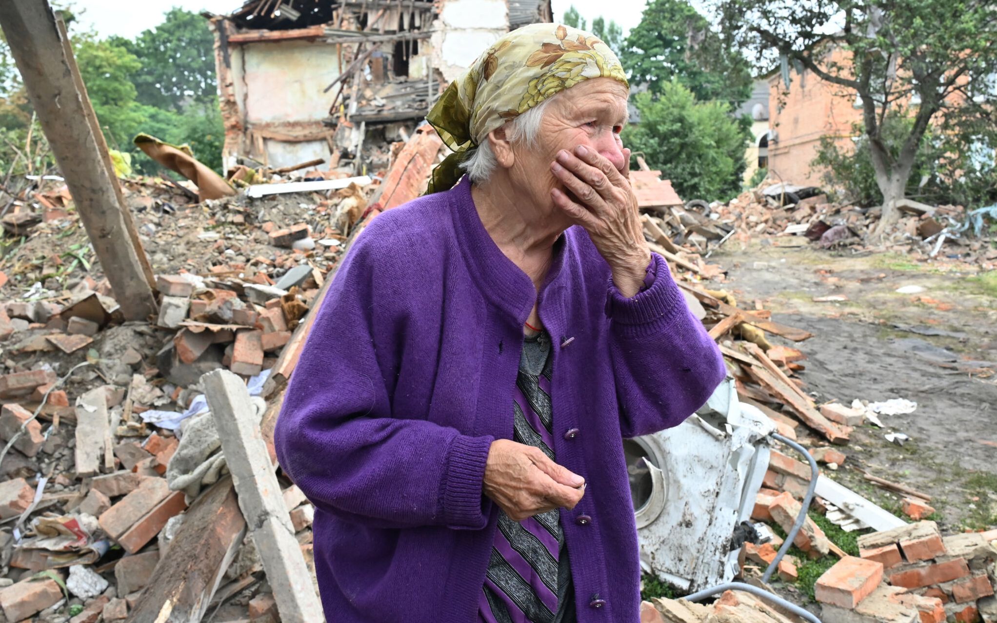 A local resident, Raisa Kuval, 82, reacts next to a damaged building partially destroyed after a shelling in the city of Chuguiv, east of Kharkiv, on July 16, 2022. - In the northeast region around Ukraine's second city of Kharkiv, governor Oleg Synegubov said an overnight Russian missile attack killed three people in the town of Chuguiv. (Photo by SERGEY BOBOK / AFP)