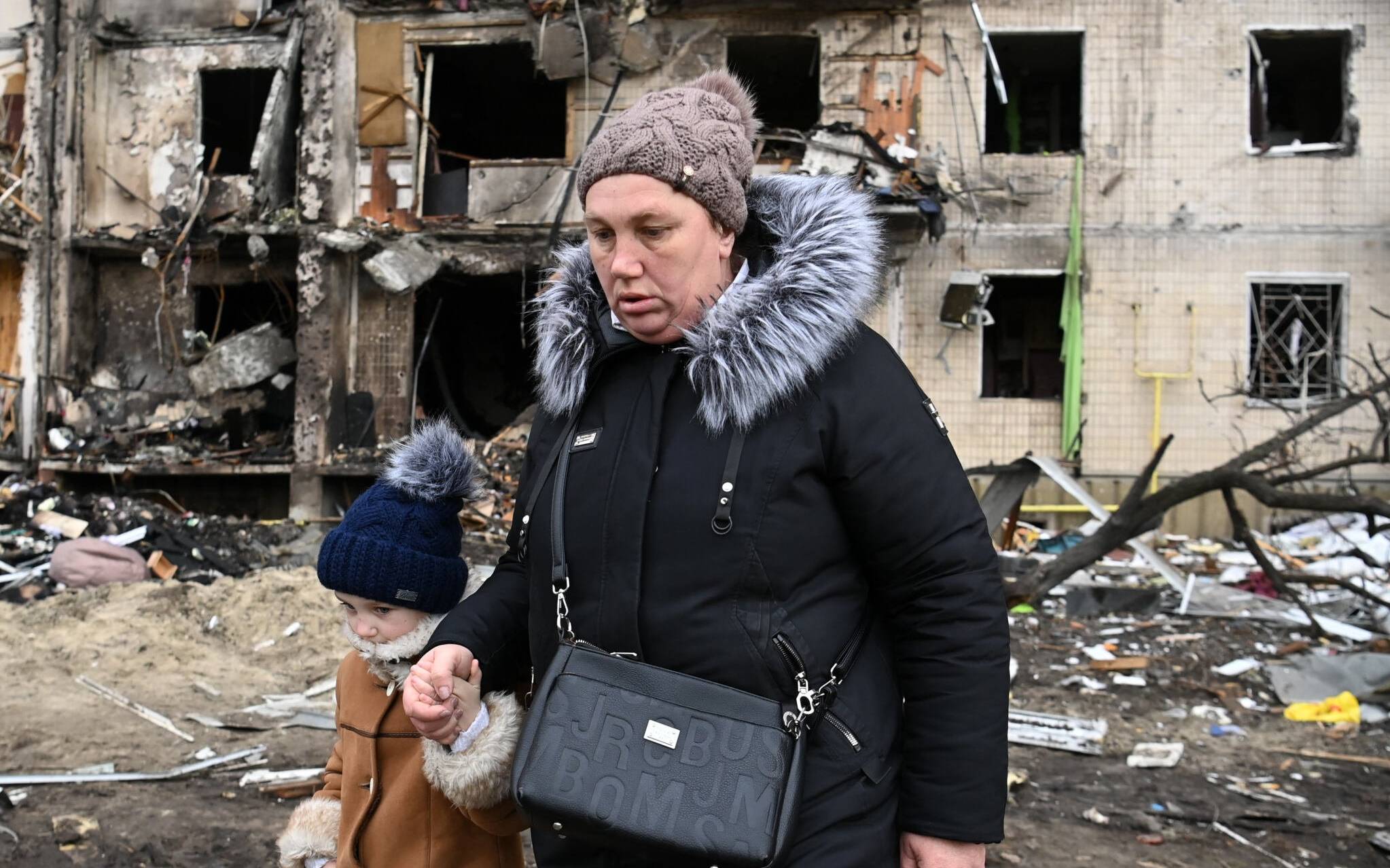 A woman with a child walks in front of a damaged residential building at Koshytsa Street, a suburb of the Ukrainian capital Kyiv, where a military shell allegedly hit, on February 25, 2022. - Russian forces reached the outskirts of Kyiv on Friday as Ukrainian President Volodymyr Zelensky said the invading troops were targeting civilians and explosions could be heard in the besieged capital. Pre-dawn blasts in Kyiv set off a second day of violence after Russian President Vladimir Putin defied Western warnings to unleash a full-scale ground invasion and air assault on Thursday that quickly claimed dozens of lives and displaced at least 100,000 people. (Photo by Daniel LEAL / AFP)