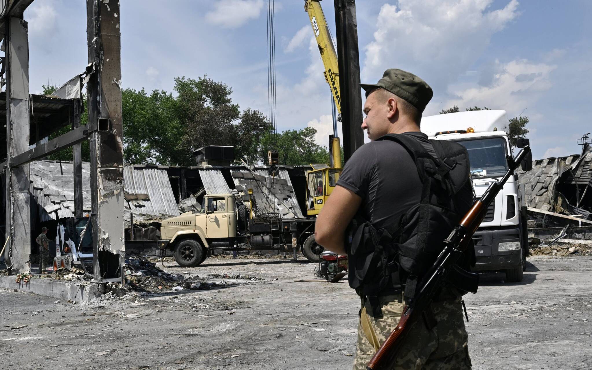 A Ukrainian serviceman stands alert as workers clear rubble and debris from The Amstor Mall in Kremenchuk, on June 29, 2022, two days after it was hit by a Russian missile strike according to Ukrainian authorities. - A missile strike on a crowded mall in Kremenchuk, central Ukraine killed at least 18 people. (Photo by Genya SAVILOV / AFP)
