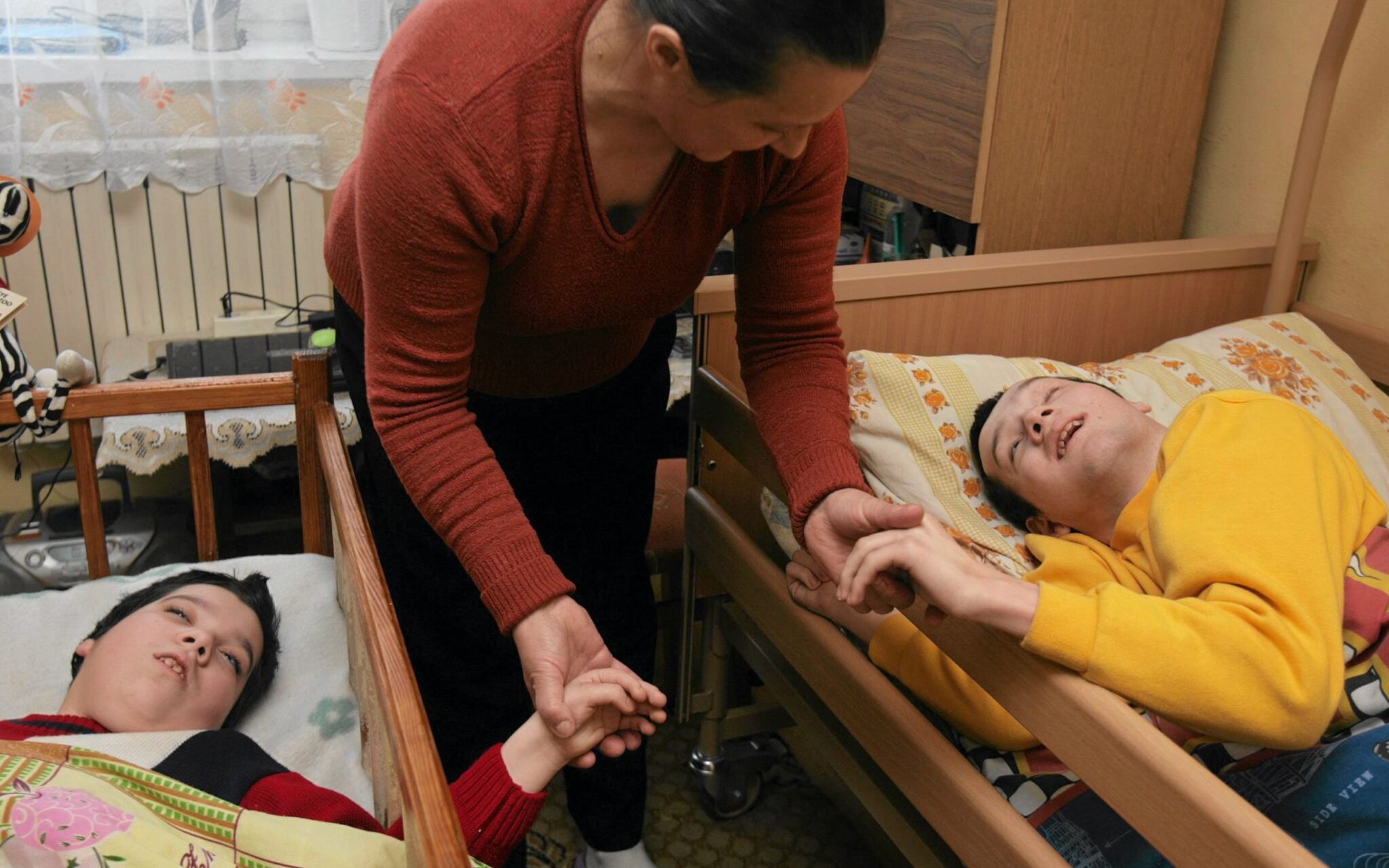 Duże dzieci z niepełnosprawnością leżą w łóżkach, pochyla się nad nimi kobieta
