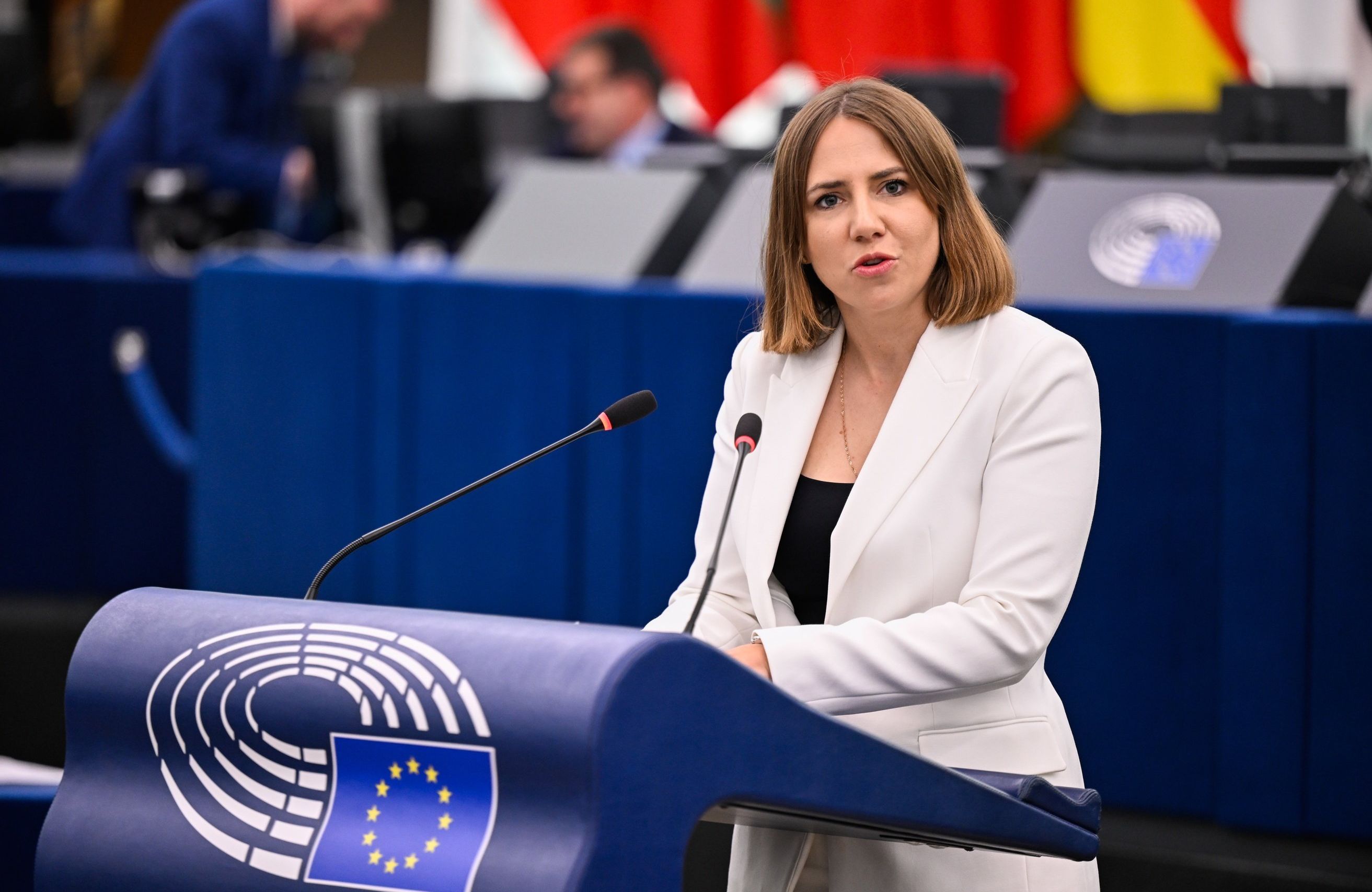 Młoda kobieta z krótkimi brązowymi włosami w jasnej marynarce i czarnej bluzce przemawia z mównicy Parlamentu Europejskiego.