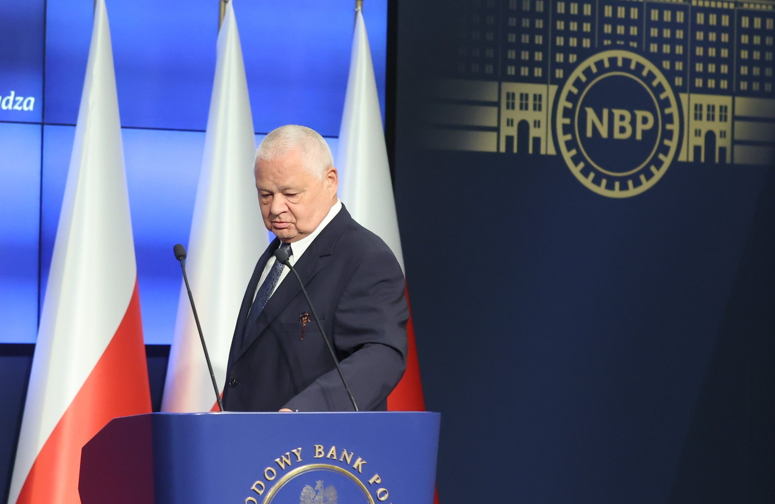 Siwy mężczyzna w garniturze schodzi z mównicy, w tle flagi Polski i logo NBP