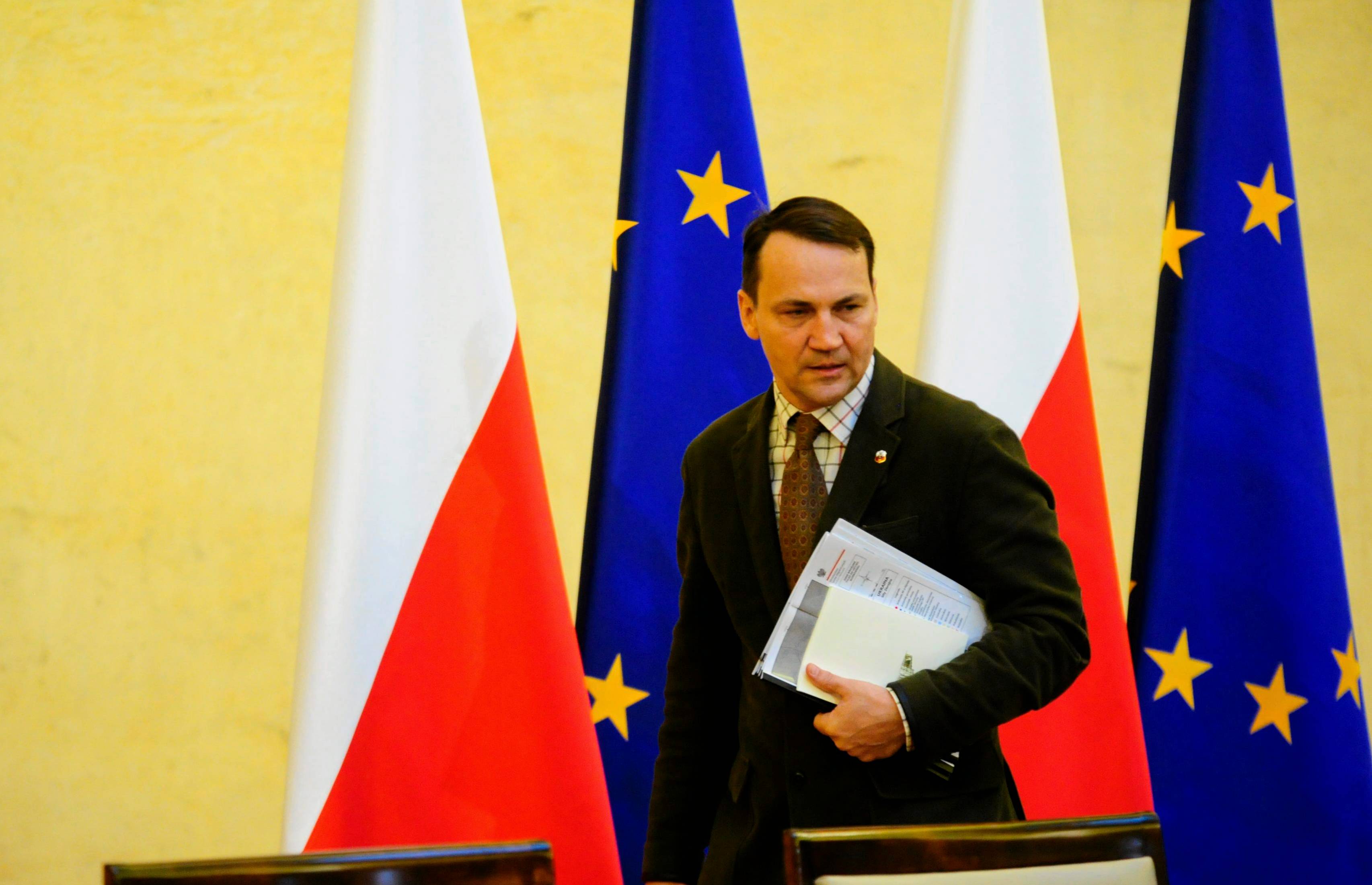 Radosław Sikorski z dokumentami na tle polskich i unijnych flag (żółta ściana)