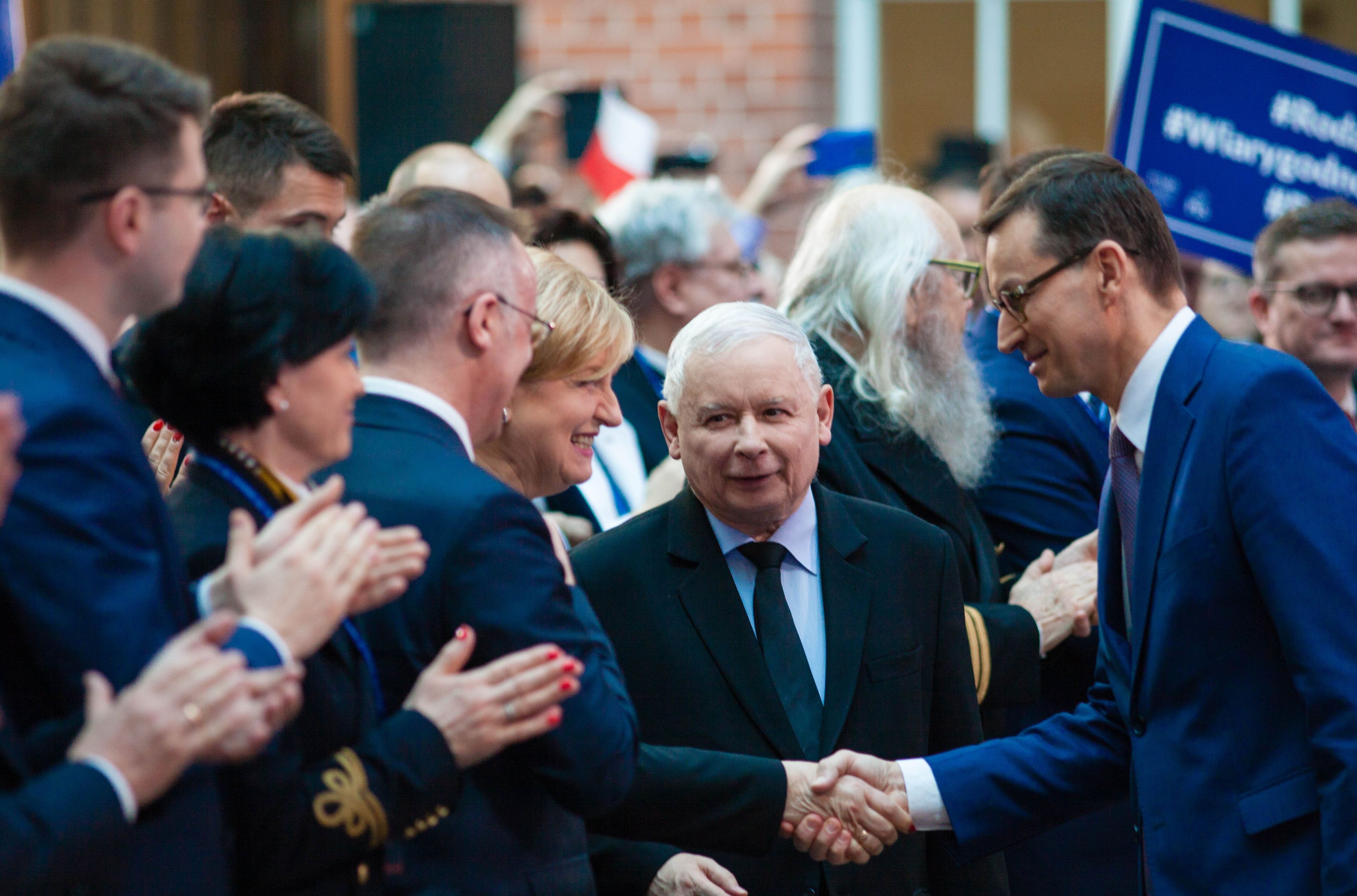 Wyższy mężczyzna z prawej ściska rękę niższego, starszego mężczyzny z lewej. Obaj są ubrani w garnitury, obok nich uśmiechnięty, wiwatujący tłum