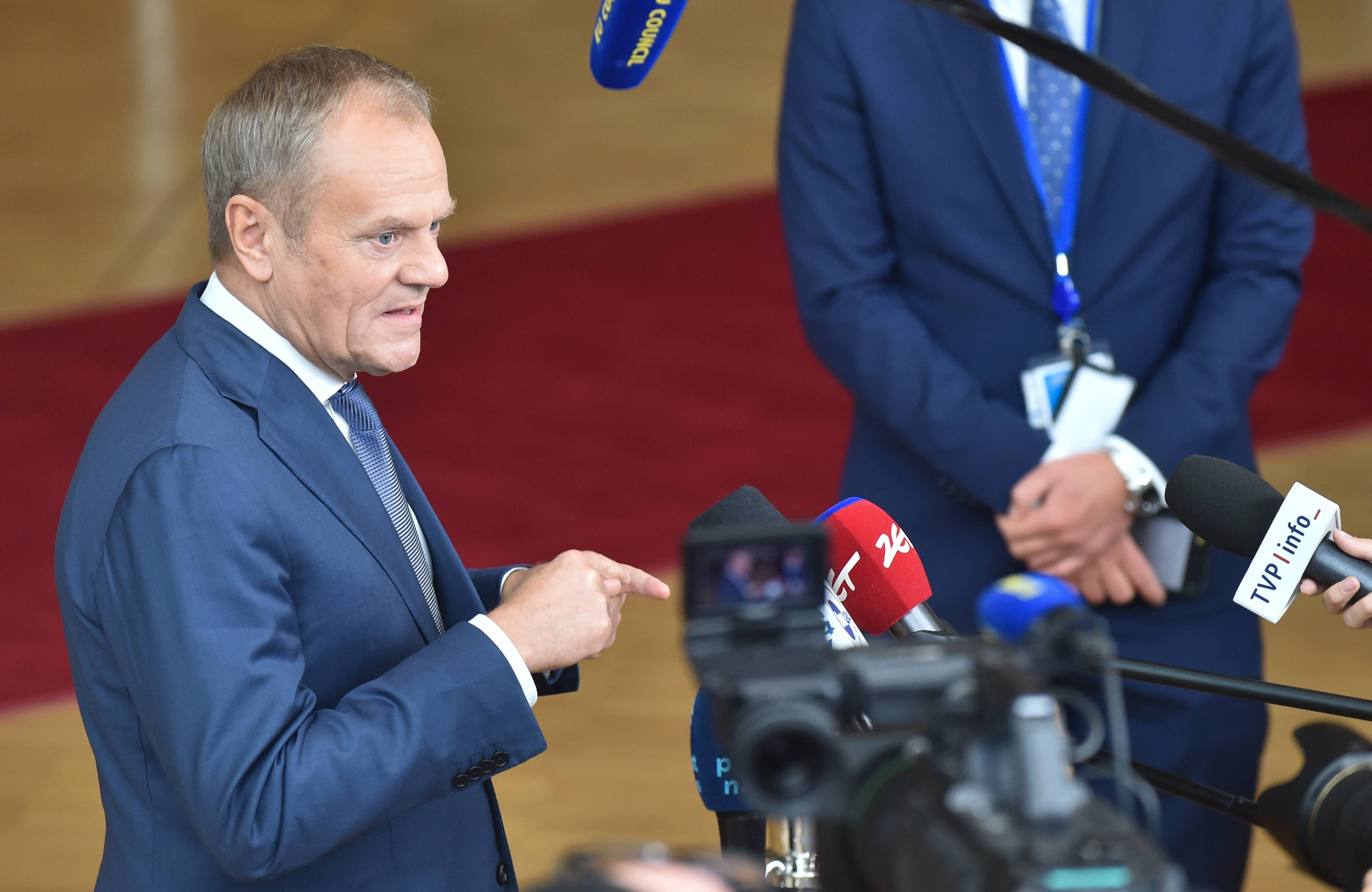 Premier Donald Tusk w niebieskim garniturze stoi przed mikrofonami dziennikarzy w holu prowadzącym do sali plenarnej budynku Rady Europejskiej, w tle widać czerwony dywan.