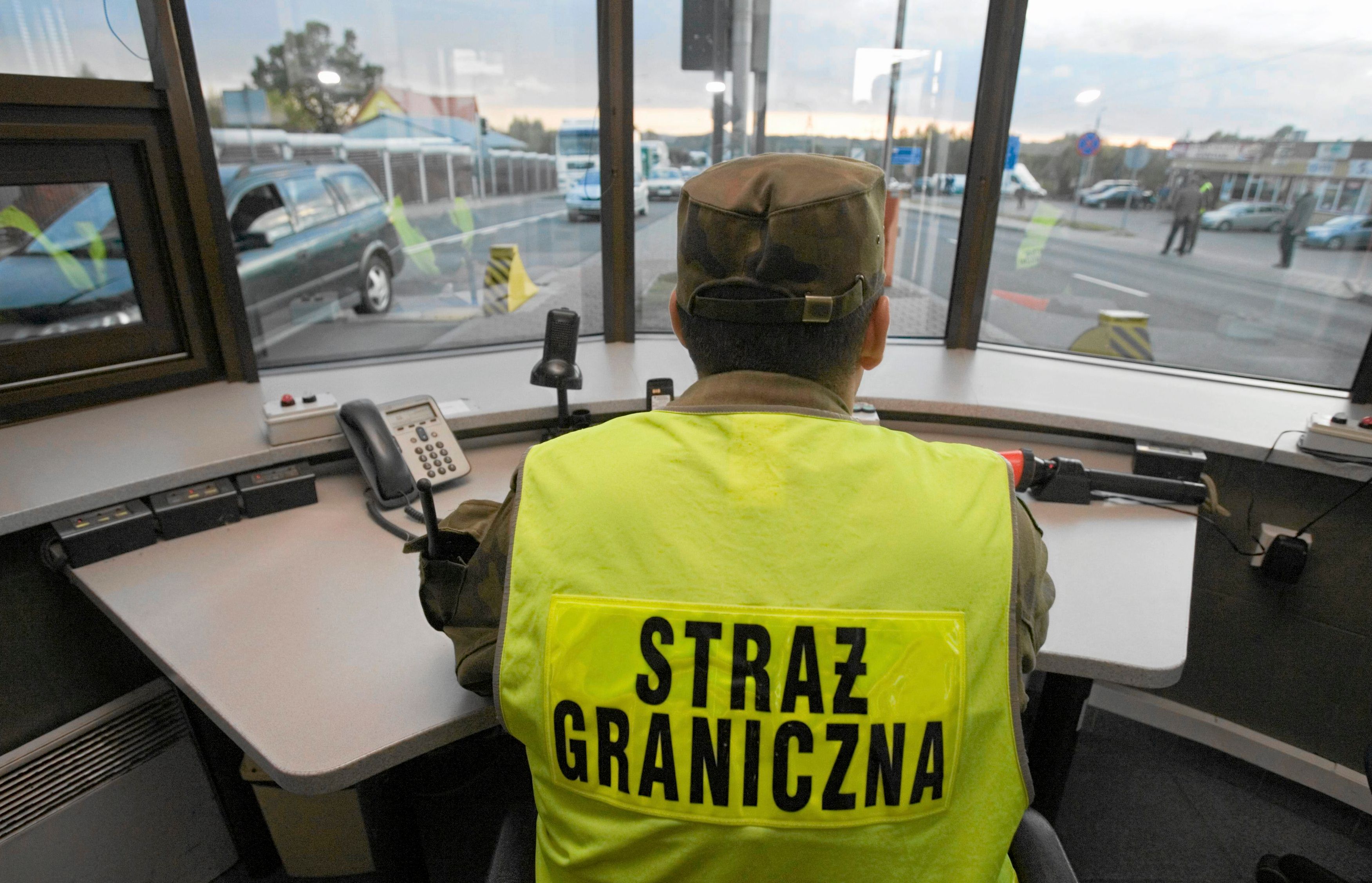 Na zdjęciu strażnik graniczny na przejściu w Kuźnicy Białostockiej. Mężczyzna w żółtej kamizelce i mundurze siedzi tyłem do obiektywu i kieruje ruchem drogowym na przejściu granicznym.
