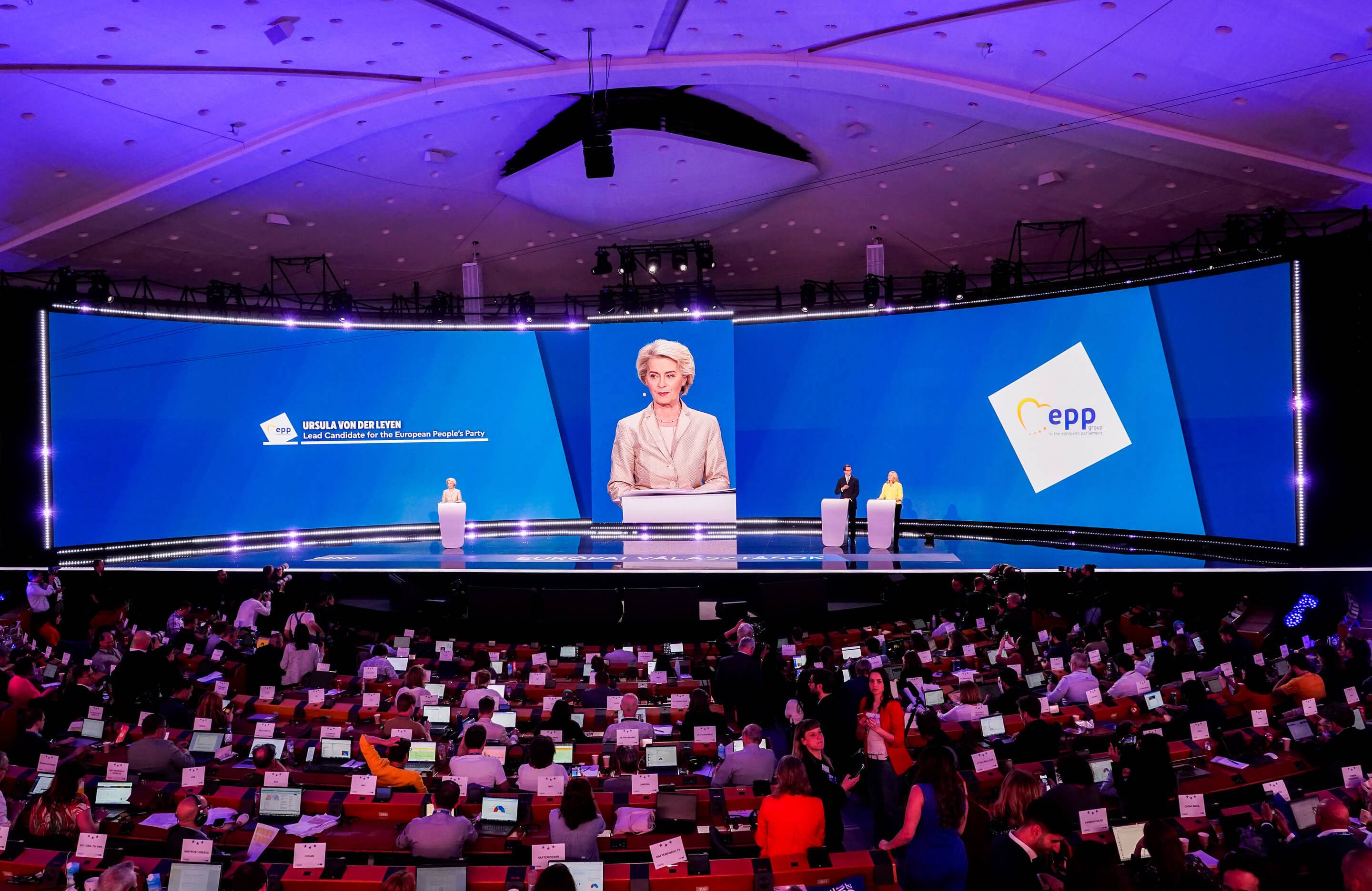 Oświetlona na niebiesko-fioletowo, pogrążona w półmroku sala plenarna Parlamentu Europejskiego, na pierwszym planie widać wiele siedzeń zajętych przez dziennikarzy, na drugim planie scena i duży ekran za sceną, widać na nim stojącą na scenie starszą kobietę z krótkimi blond włosami, brązowo-złotym makijażu, w jasnej garsonce