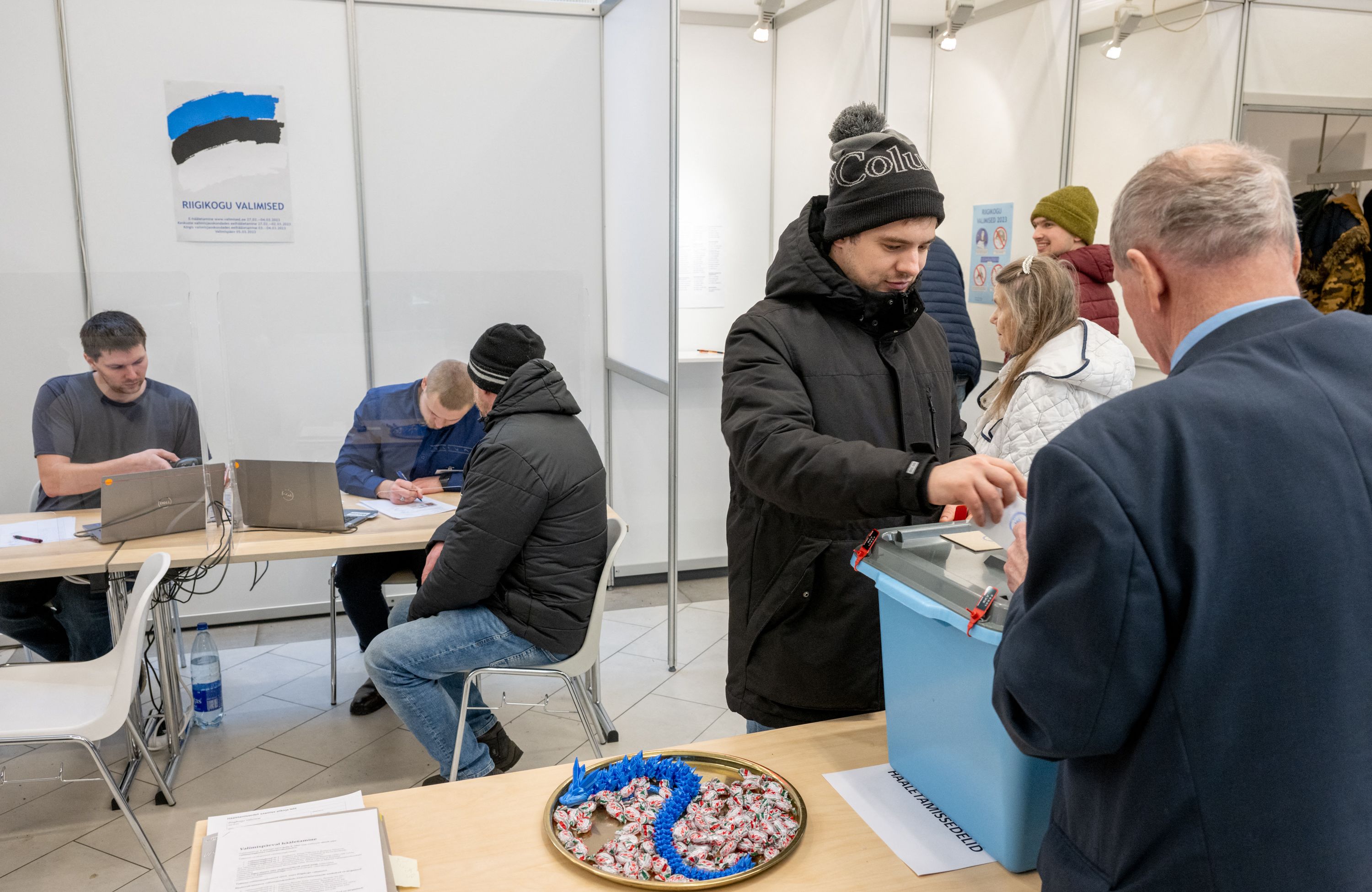 człowiek wrzuca kartę wyborcza do urny. Dwie inne osoby siedza przy stanowisku z komputerami