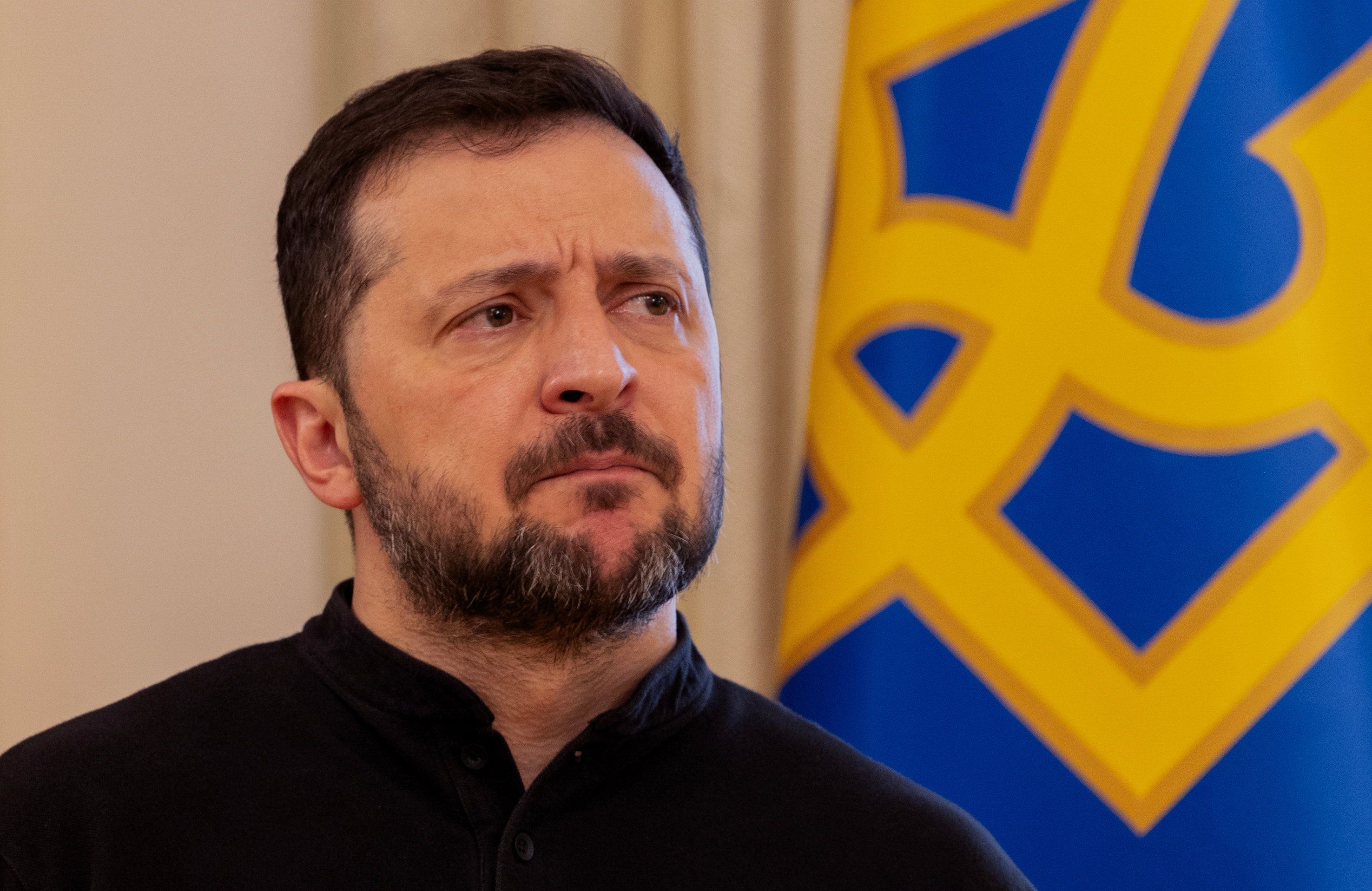 Brodaty mężczyzna (prezydent Ukrainy Wołodymyr Zełenski) siedzi na tle ukraińskiej flagi