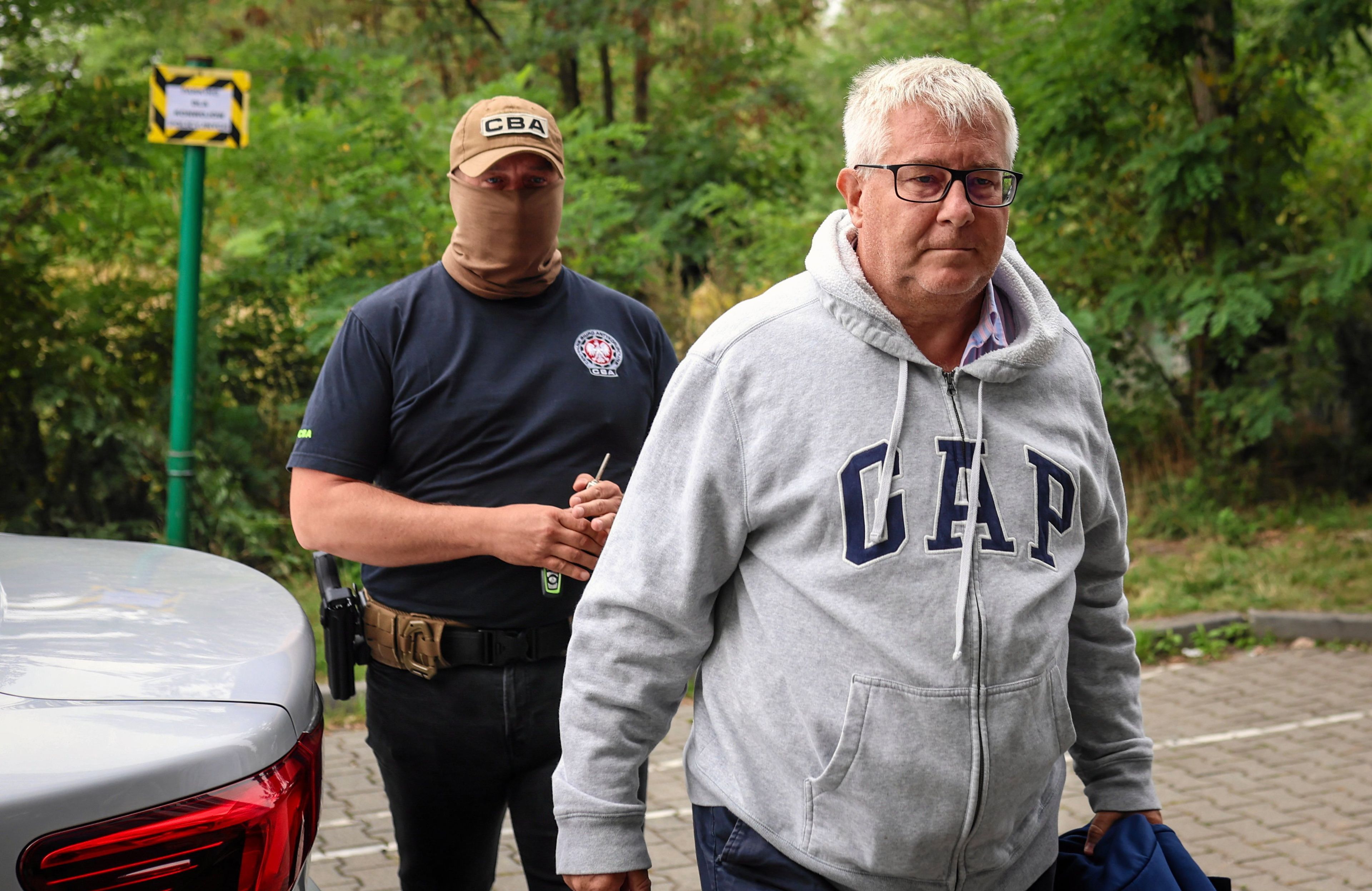 siwy korpulentny mężczyzna w okularach i w dresie GAP, za nim funkcjonariusz w mundurze