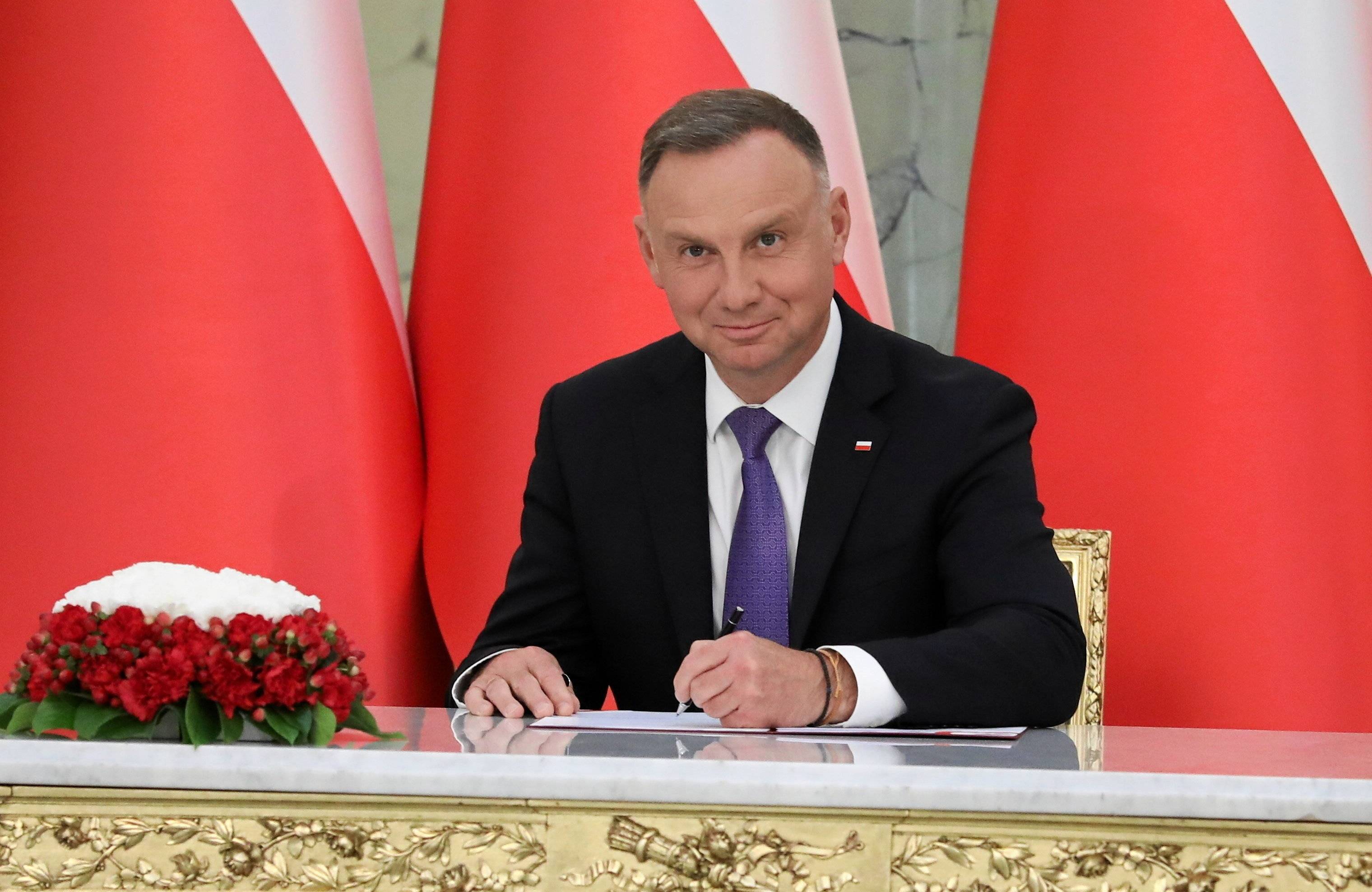 Mężczyzna w garniturze (Andrzej Duda) podpisuje dokumenty przy eleganckim stoliku. W tle biało-czerwone flagi