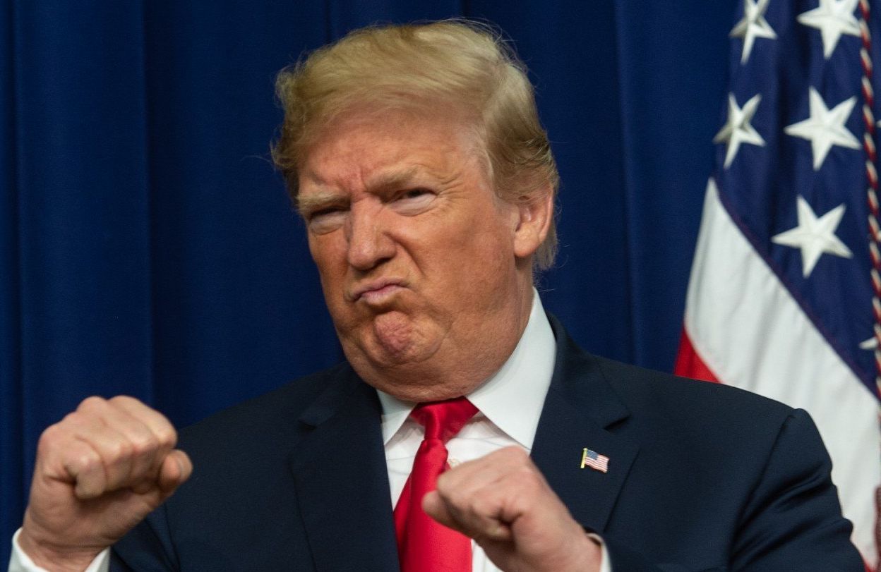 Donald Trump z pewną siebie miną i uniesionymi do góry pięściami w bokserskim geście. W tle flaga USA.
