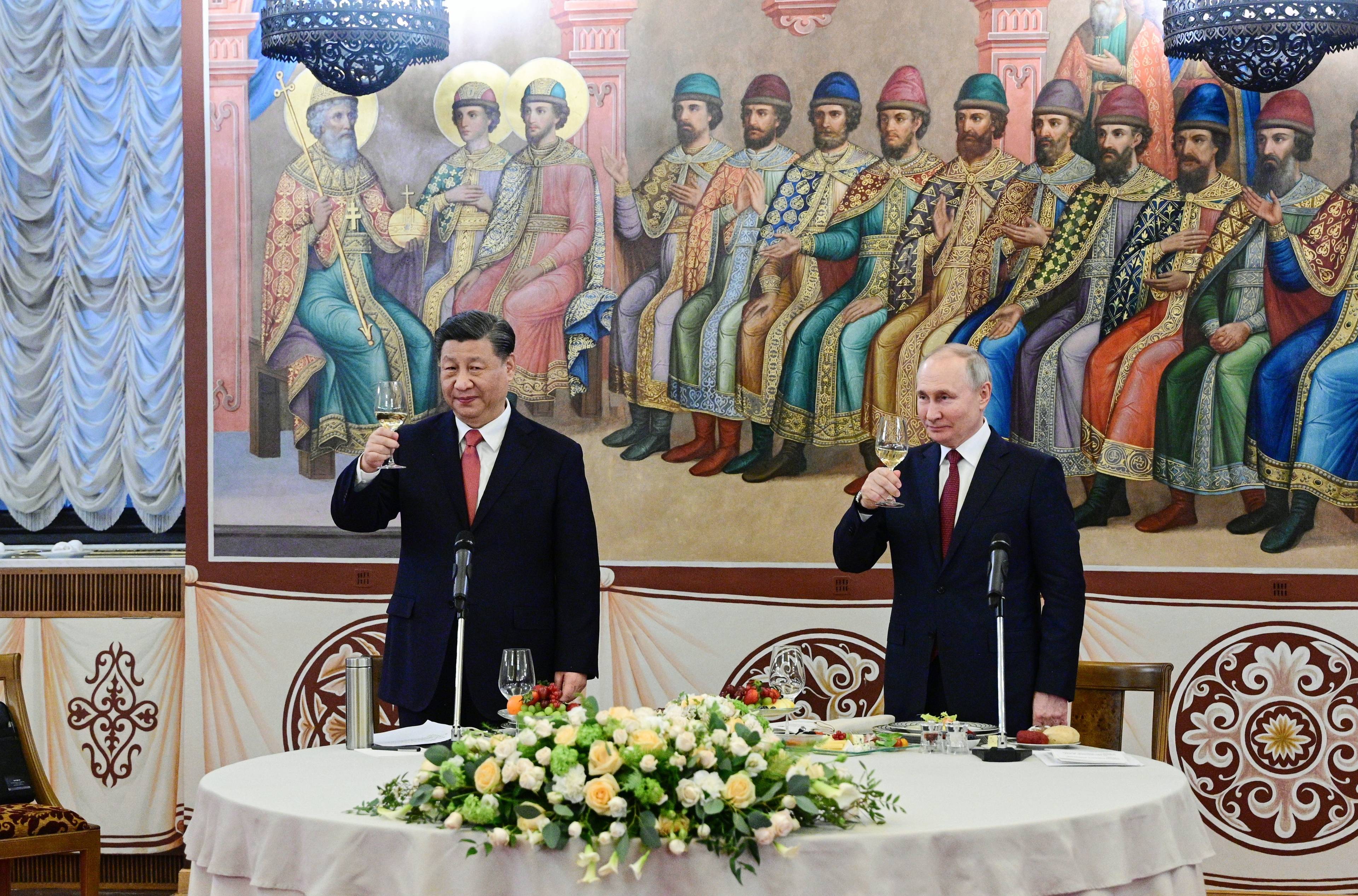 Władimir Putin i Xi Jinping stoją przy stole bankietowym z kieliszkami. Na stole żółte kwiaty, za politykami fresk