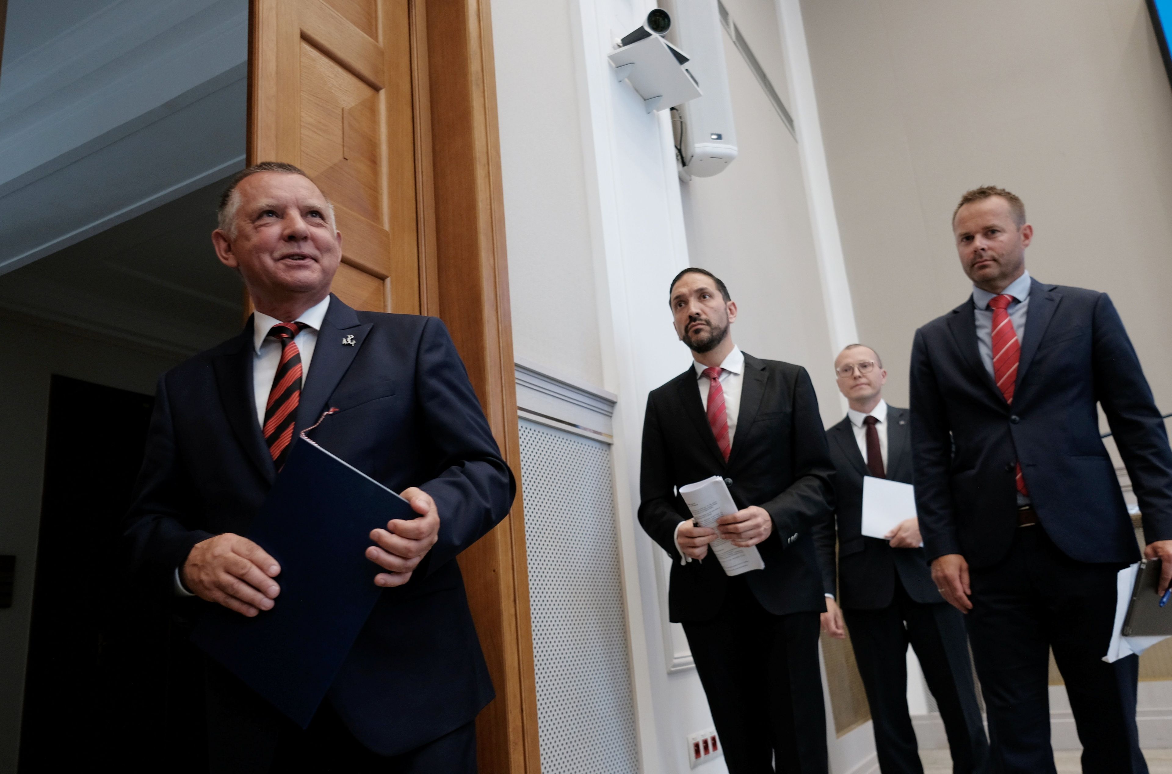 Mężczyzna z teczką (Marian Banaś prezes NIK) w drzwiach na korytarzu urzędu. Obok niego trzech innych mężczyzn w garniturach