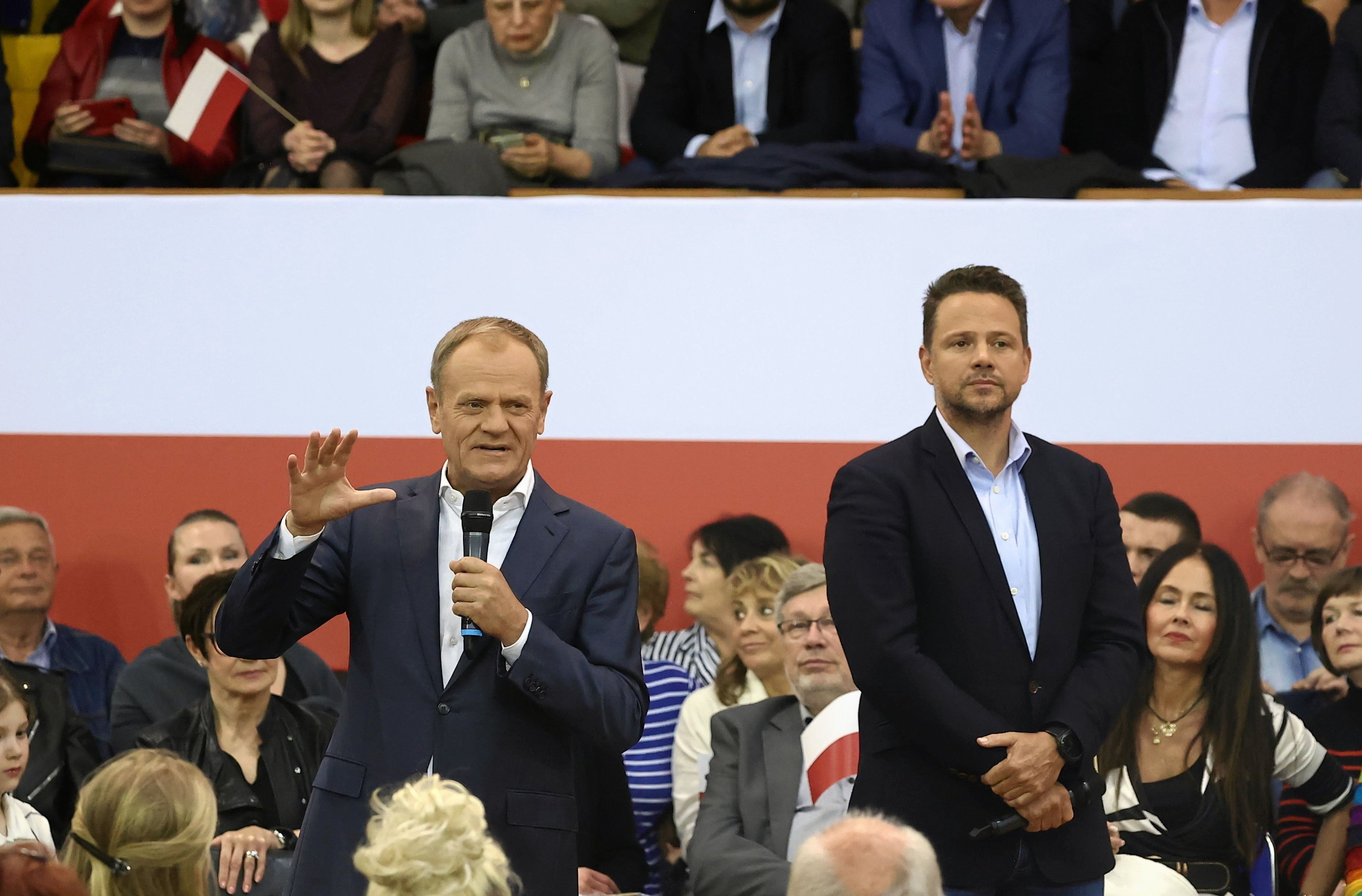 Donald Tusk przemawia na dużej sali. Za nim ludzie i flaga Polski obok niego stoi Rafał Trzaskowski. Obaj politycy są w ciemnych marynarkach i bałych koszulach, ale bez krawatów