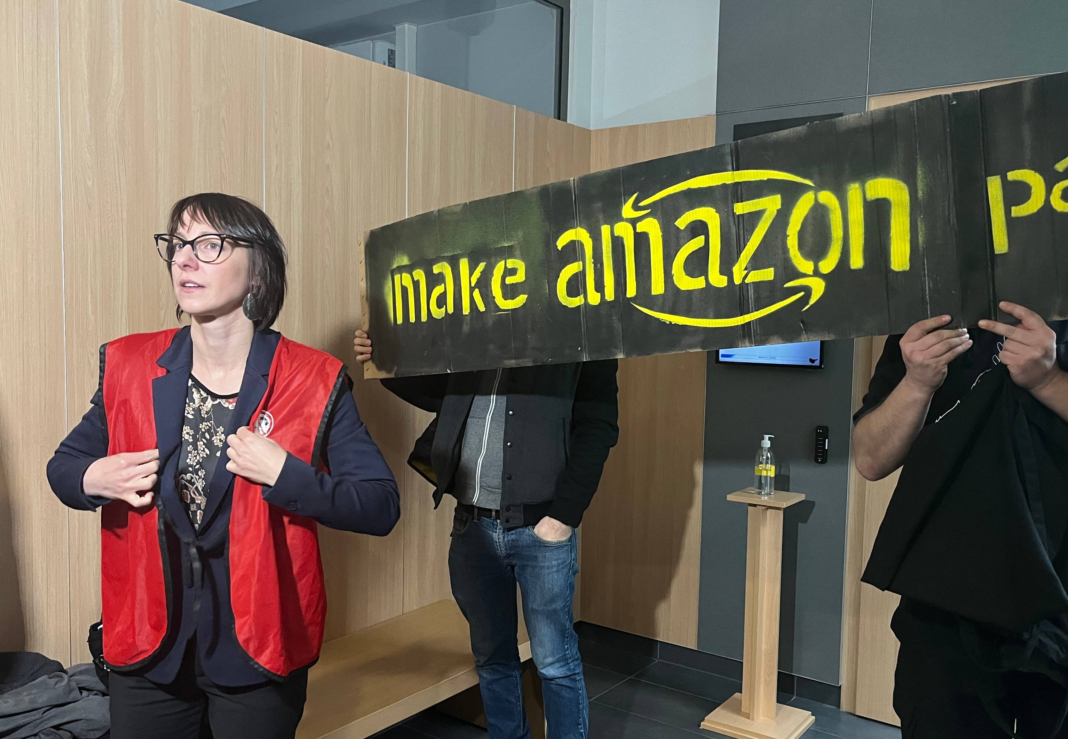 Kobieta w czerwonej kamizelce na tle transparentu "Make Amazon pay"