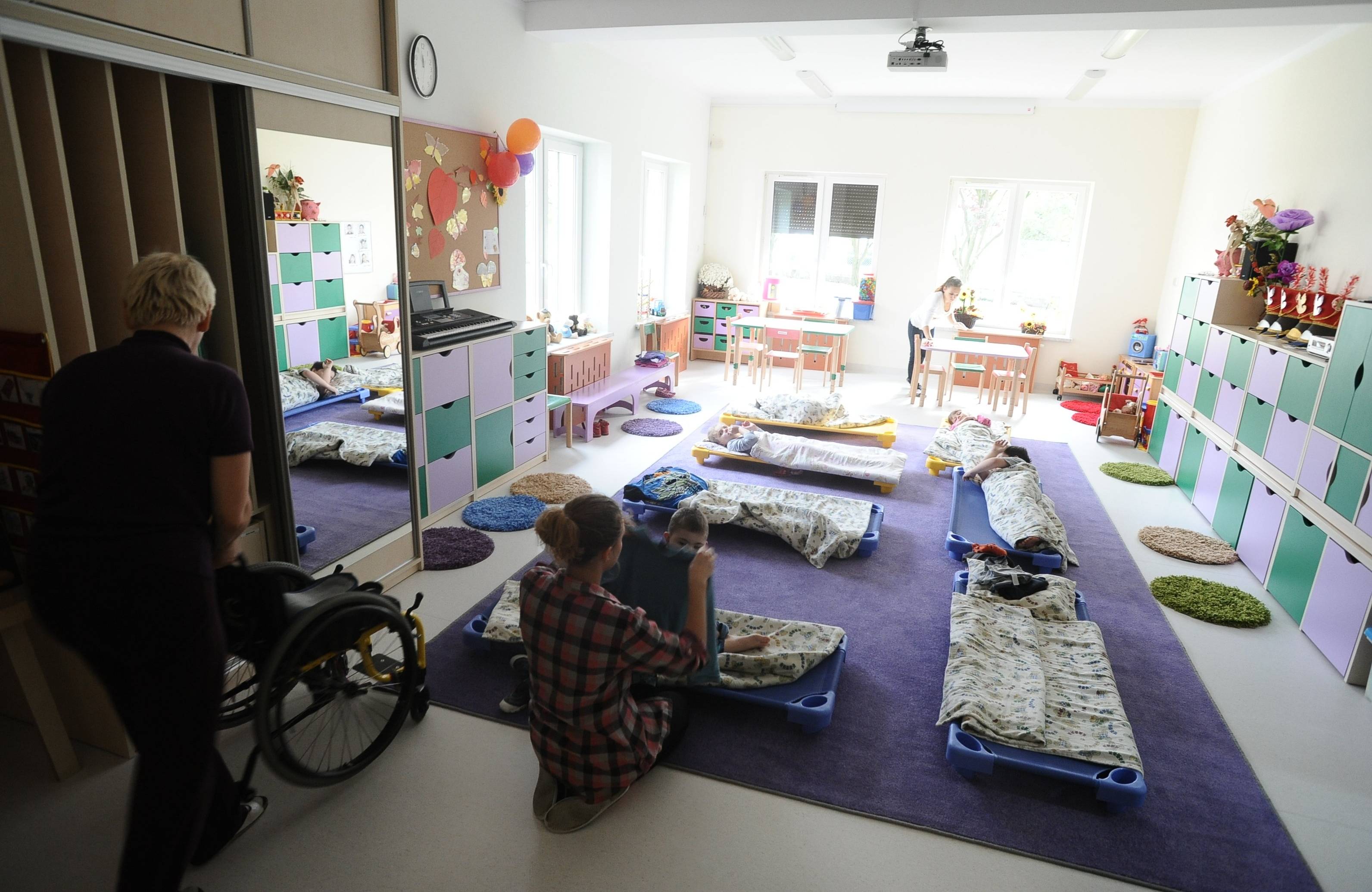 Sala przedszkolna z materacami rozłożonymi na podłodze, z boku dziecko na wózku