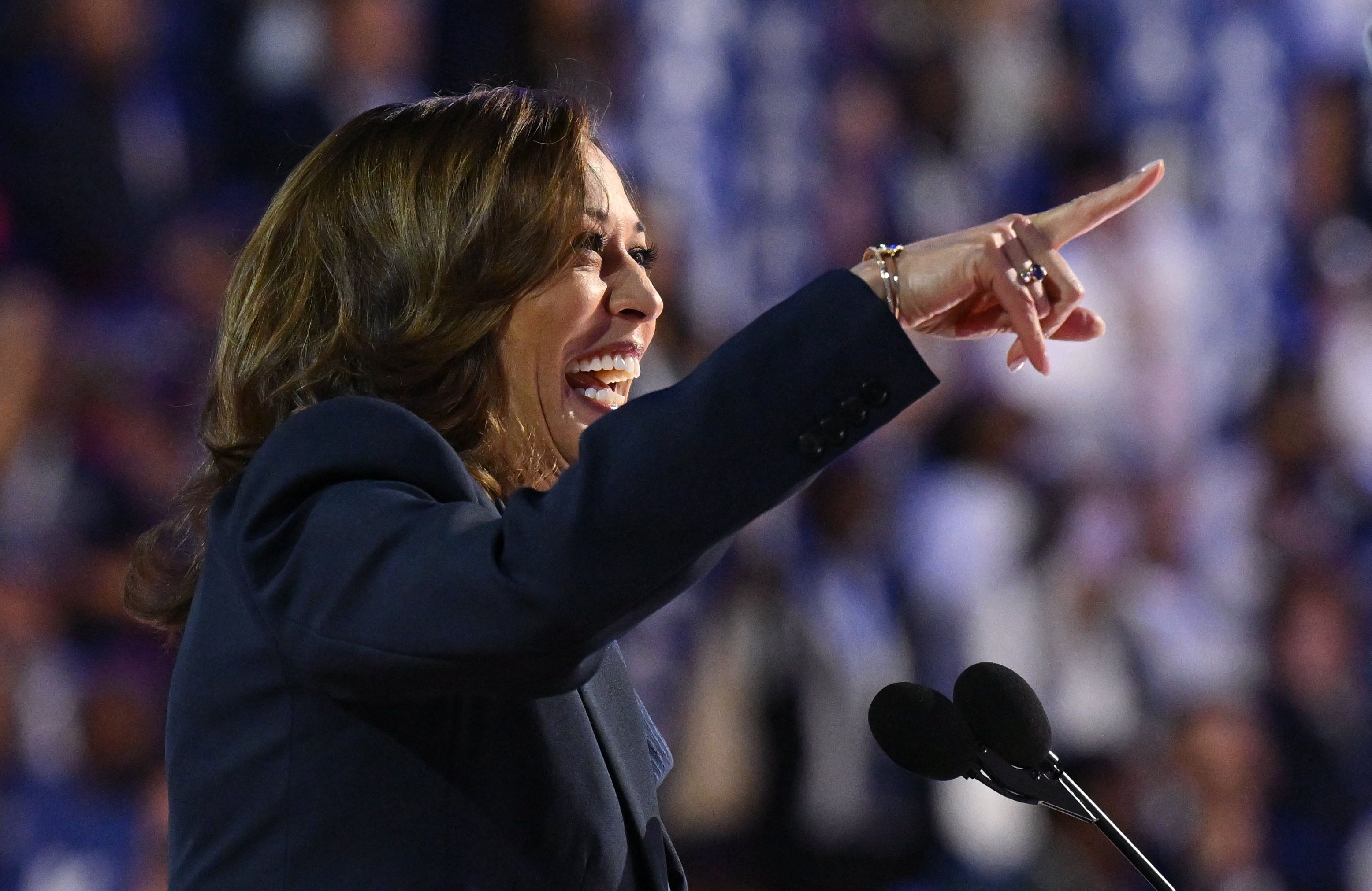 Kobieta - kamala Harris - przemawia na scenie z wyciągniętą ręką i wielkim uśmiechem na twarzy