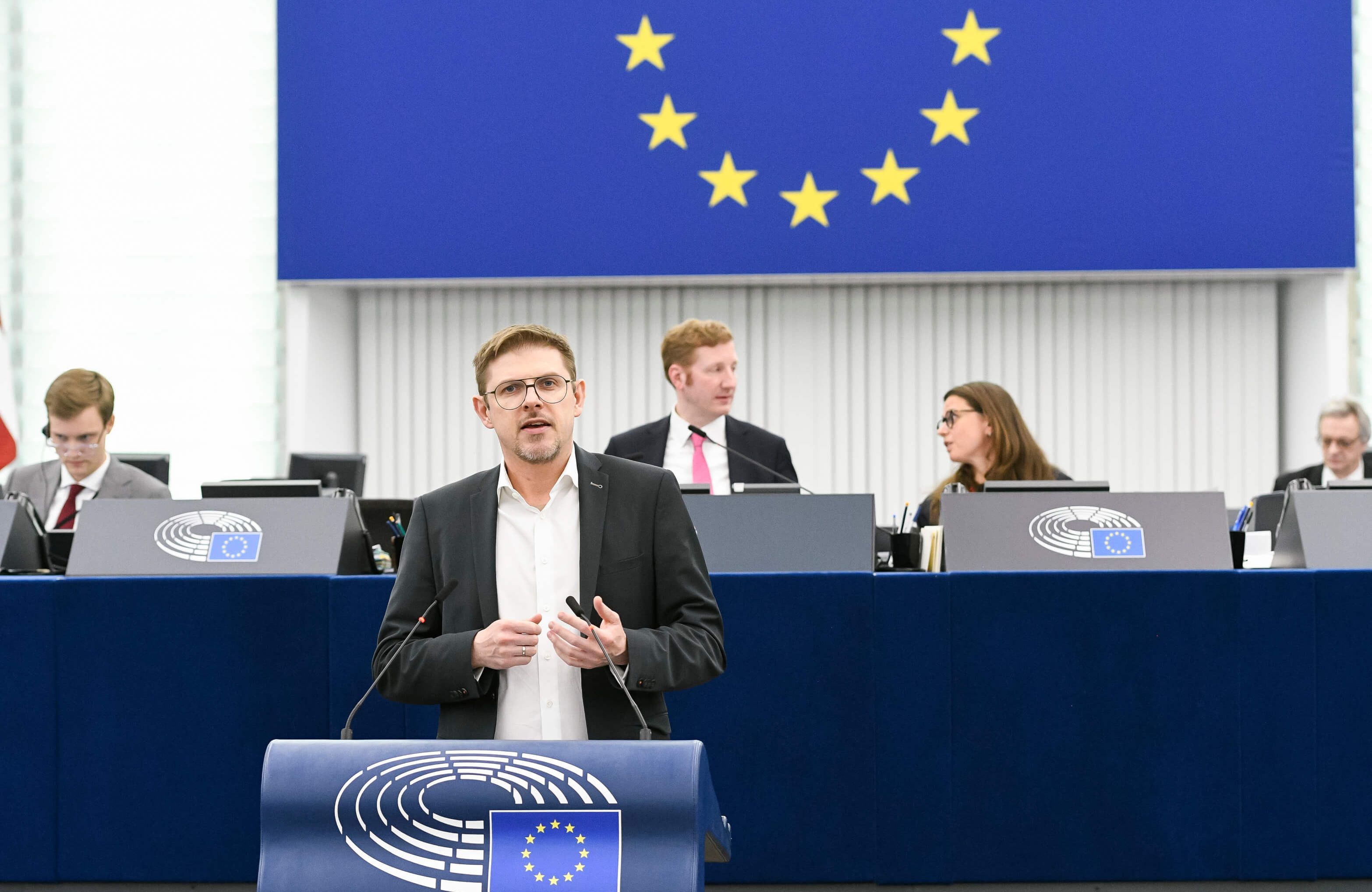 Mężczyzna z krótkimi włosami ciemny blond w okularach, białej koszuli i czarnej, casualowej marynarce stoi przy mównicy w sali plenarnej Parlamentu Europejskiego, w tle widać kawałek unijnej flagi oraz dwie osoby - kobietę i mężczyznę - innych europosłów