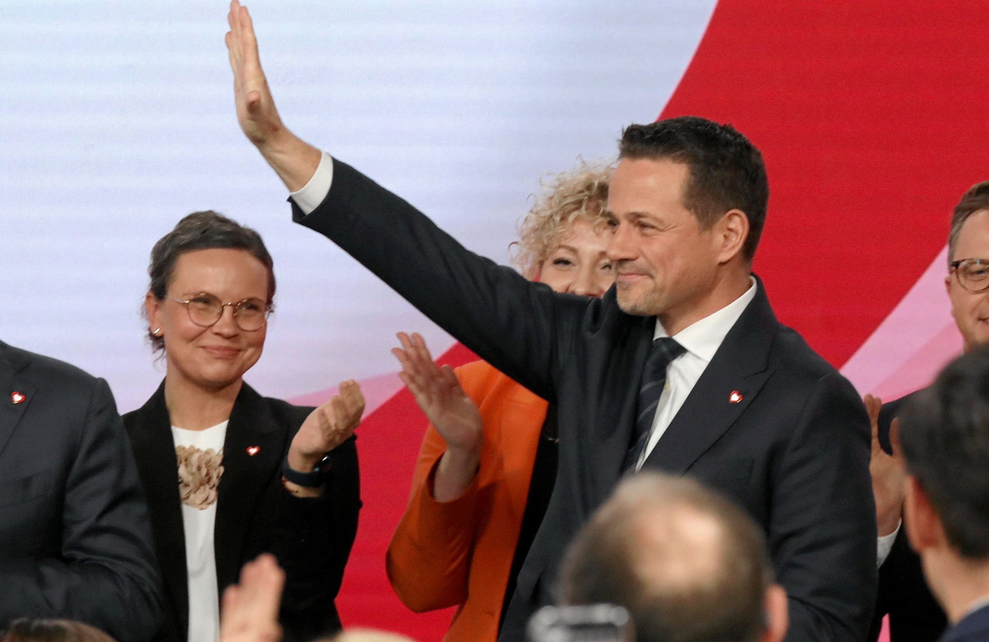 Mężczyzna w sile wieku, w garniturze, podnosi rękę w geście pozdrowienia, z tyłu osoby klaszczące