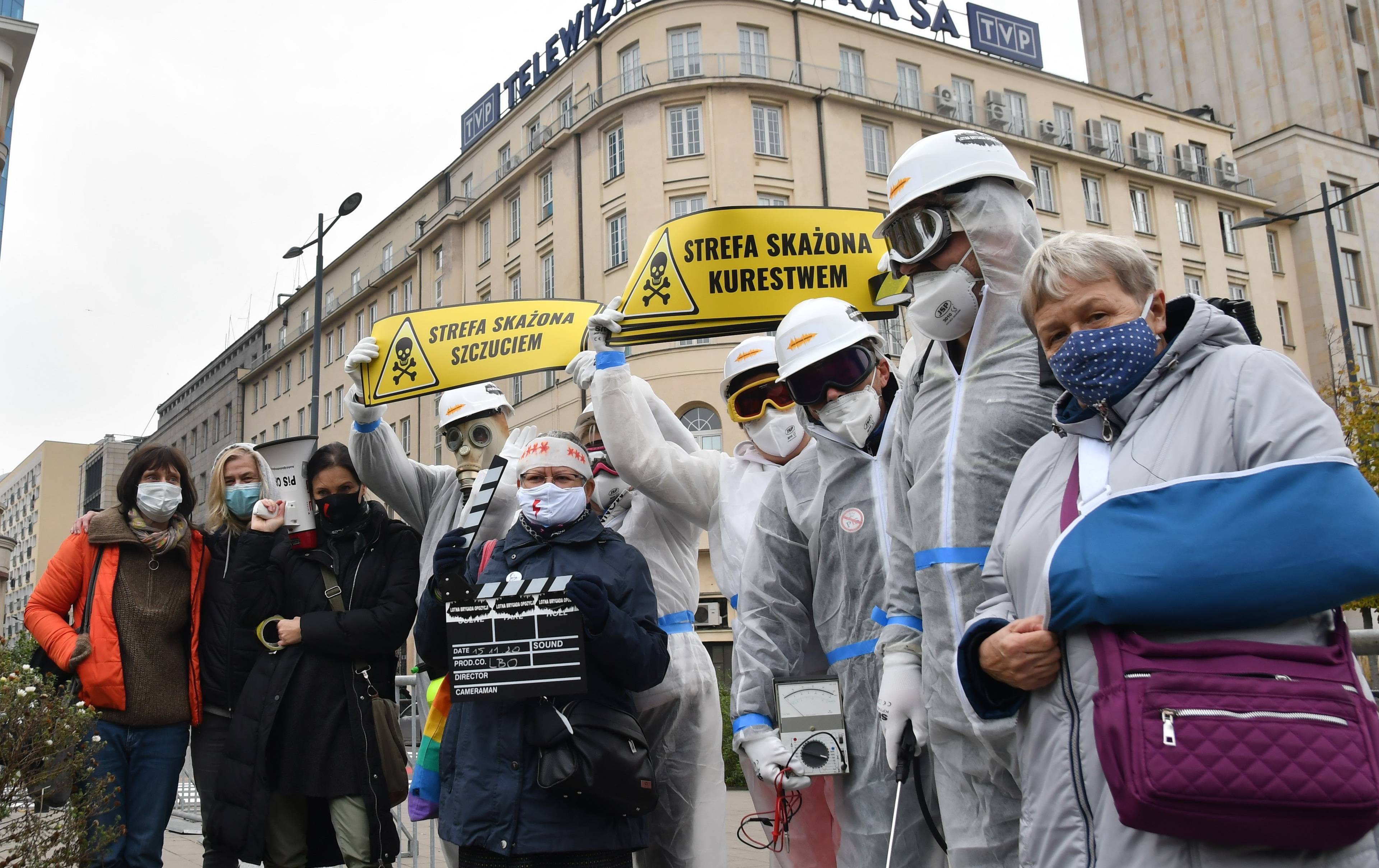Lotna Brygada Opozycji pod TVP.Aktywiści w maskach przeciwgazowych i w kombinezonach stoją pod budynkiem TVP, za nimi żółte tabliczki ostrzegawcze
