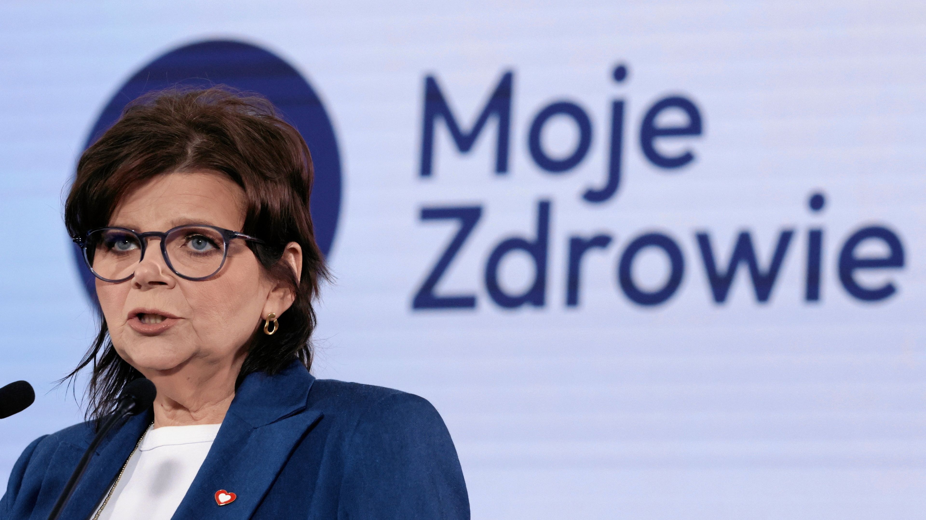 Kobieta w okulrach, w granatowej marynarce na tle planszy z napisem Moje Zdrowie