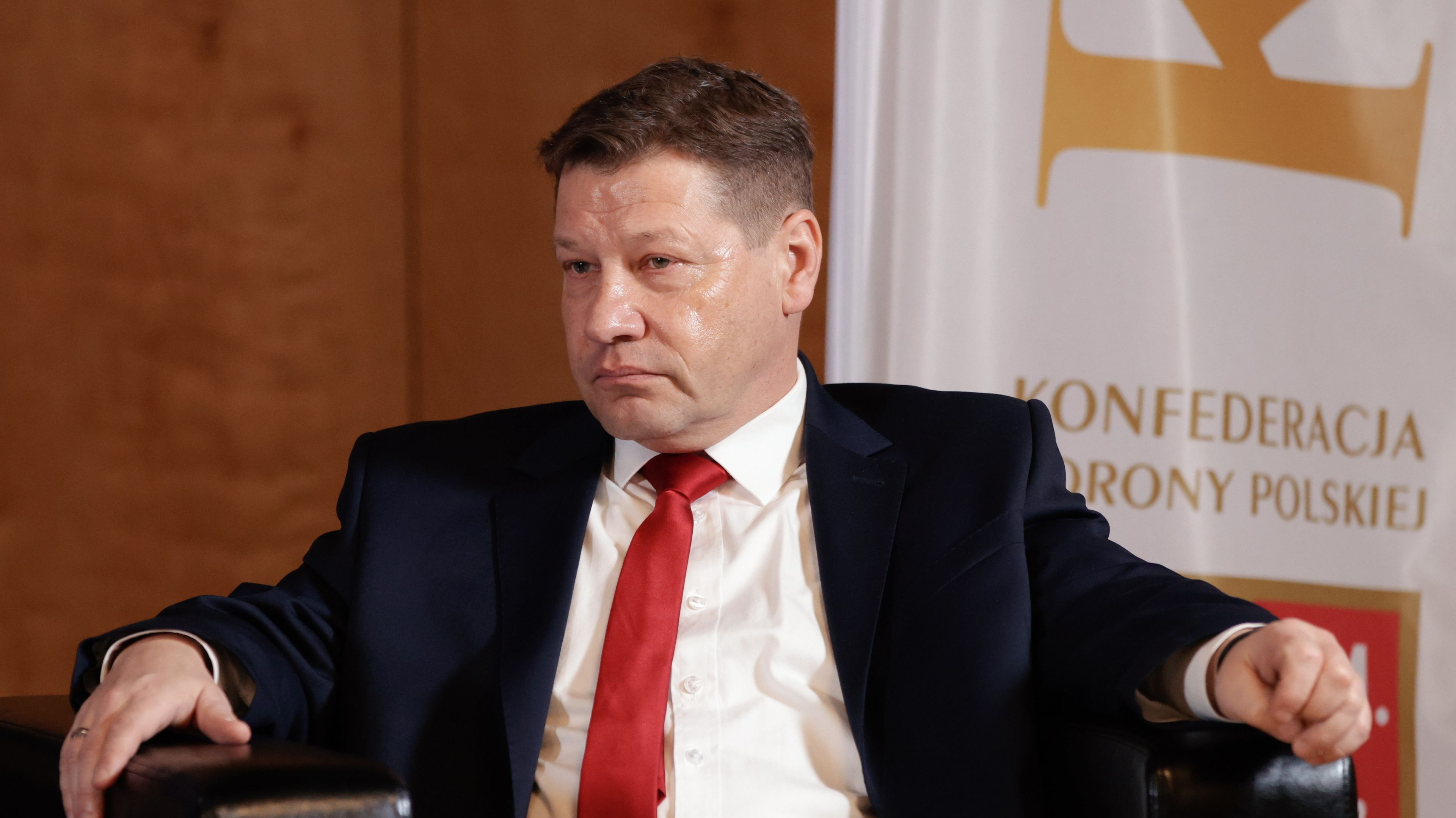 Mężczyzna w średnim wieku siedzi rozparty, w rozpiętej marynarce, ma czerwony krawat do białej koszuli.