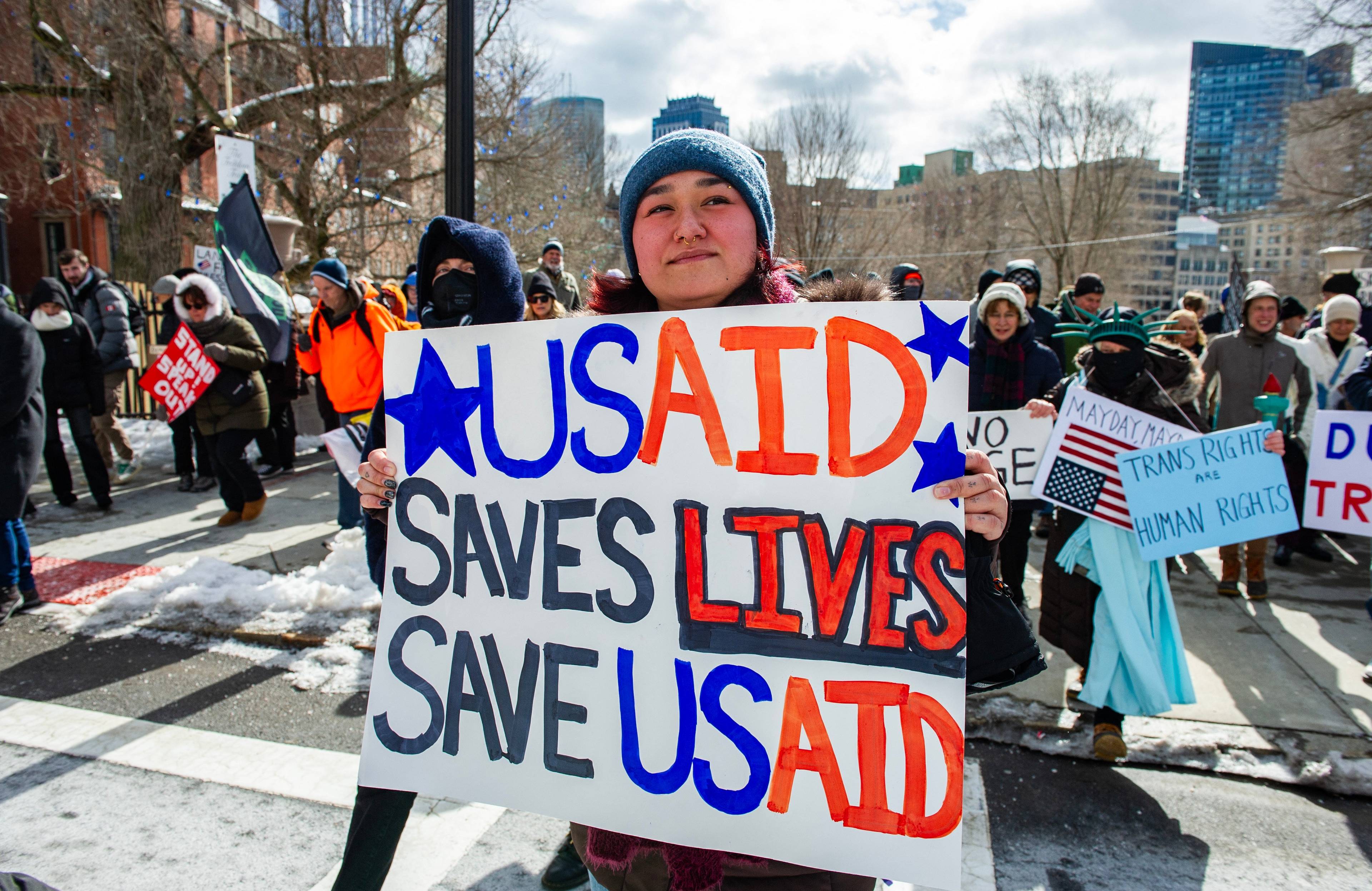 Młoda kobieta w zimowej czapce uczestniczy w ulicznym proteście, trzymając transparent z napisem "USAID saves lives. Save USAID"