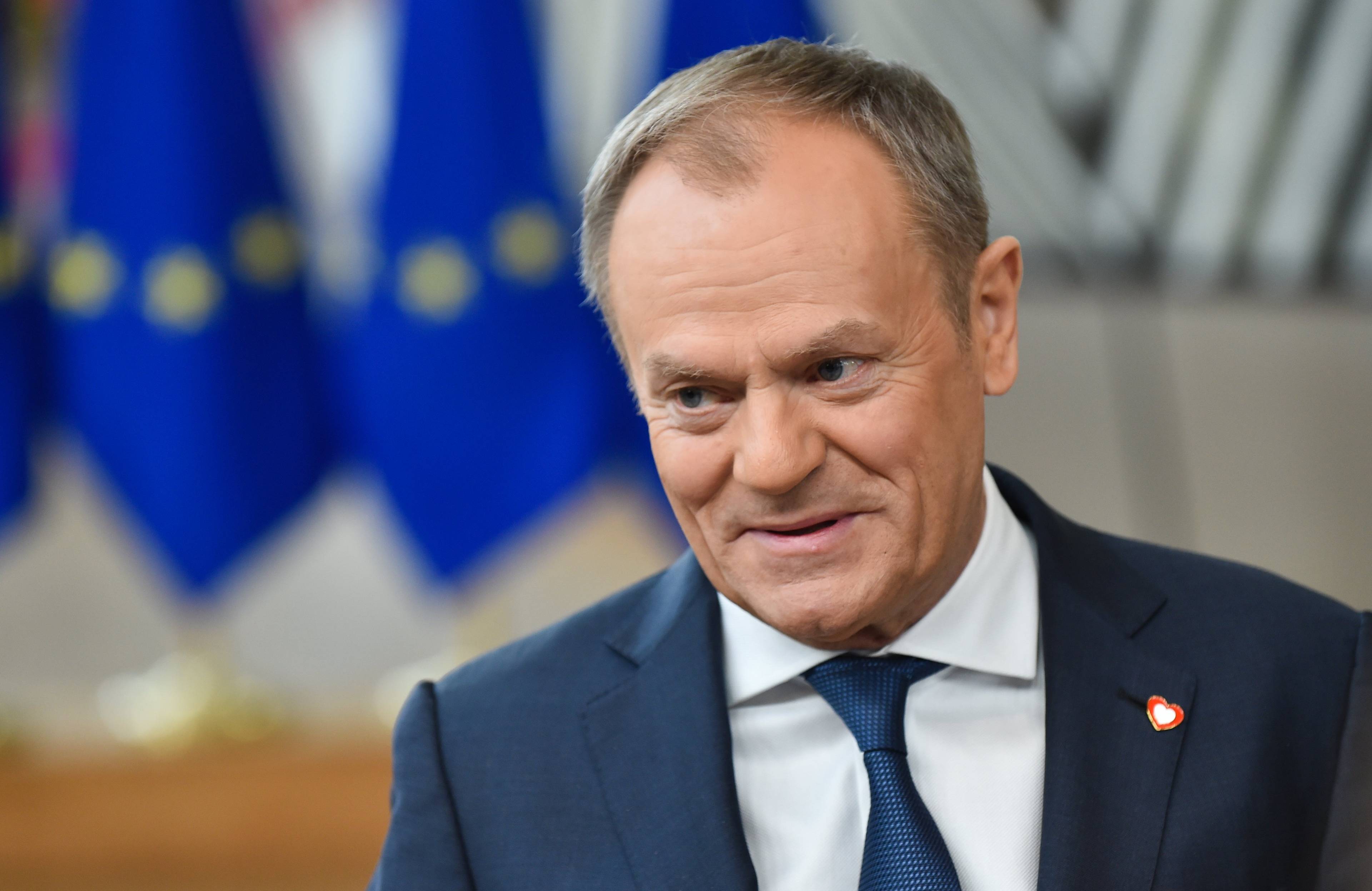 Premier Polski Donald Tusk w niebieskim garniturze i niebieskim krawacie uśmiecha się rozmawiając z dziennikarzami tuż przed rozpoczęciem szczytu Unii Europejskiej w lutym 2024; w tle niebieska flaga UE z żółtymi gwiazdkami