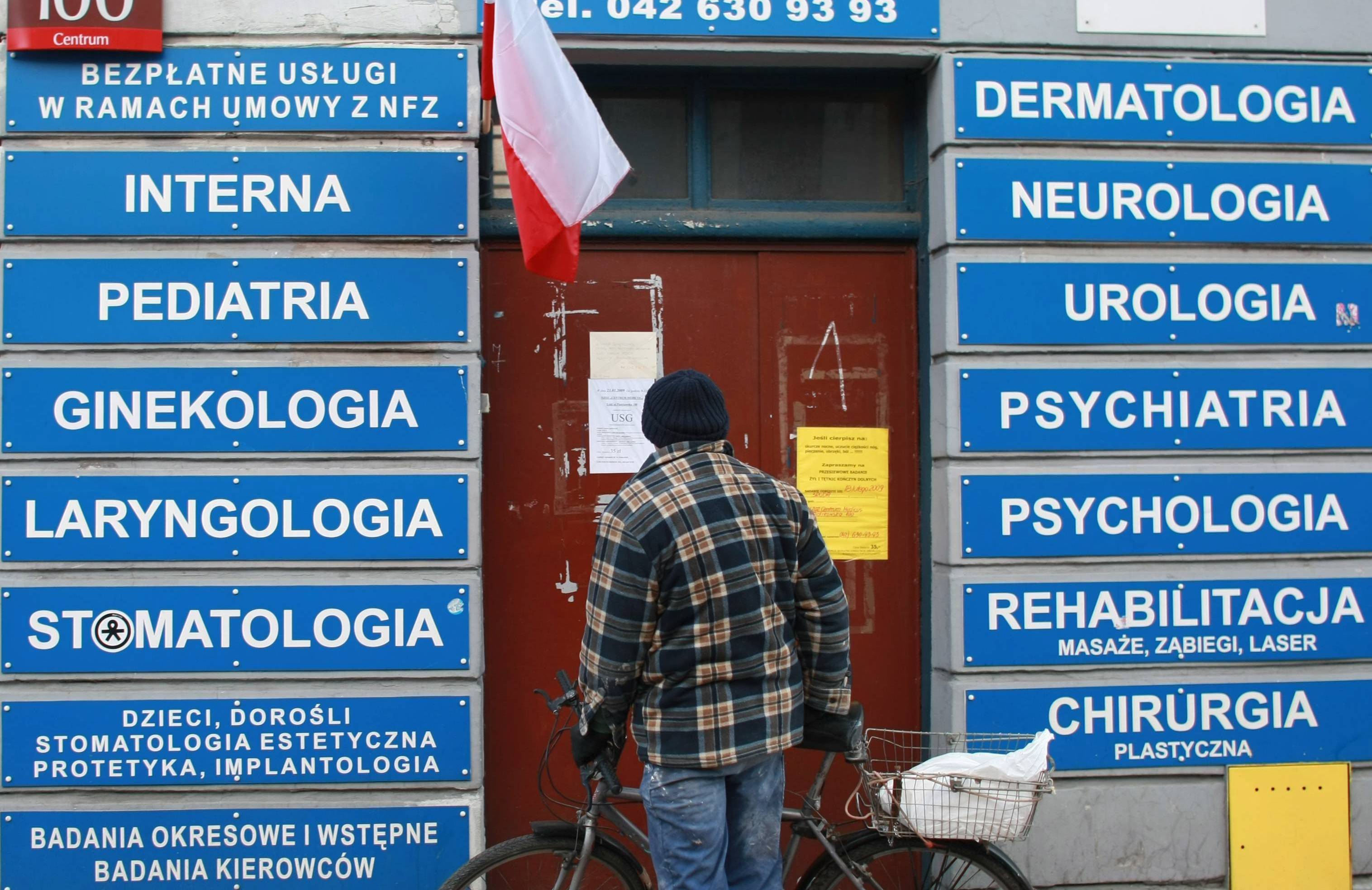 26.01.2009, Łódź. Mężczyzna w kraciastej koszuli stoi z rowerem przed wejściem do przychodni. Po obu stronach drzwi wiszą niebieskie szyldy z nazwami różnych specjalizacji: "Interna", "Pediatria", "Laryngologia". Nad drzwiami niebieski szyld z napisem "Przychodnia". Obok logo NFZ i wywieszona polska flaga