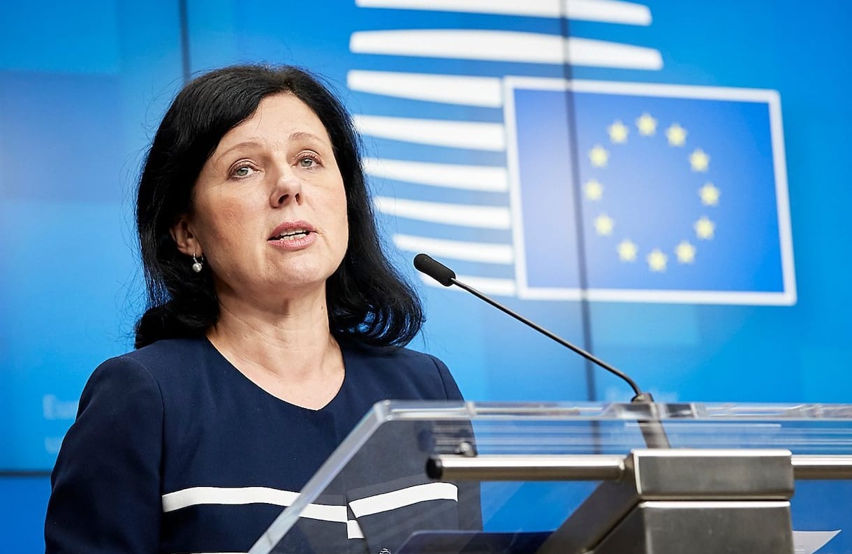 40-letnia brunetka z włosami do ramion przemawia znad pulpitu, w tle flaga UE - gwiazdy na niebieskim tle