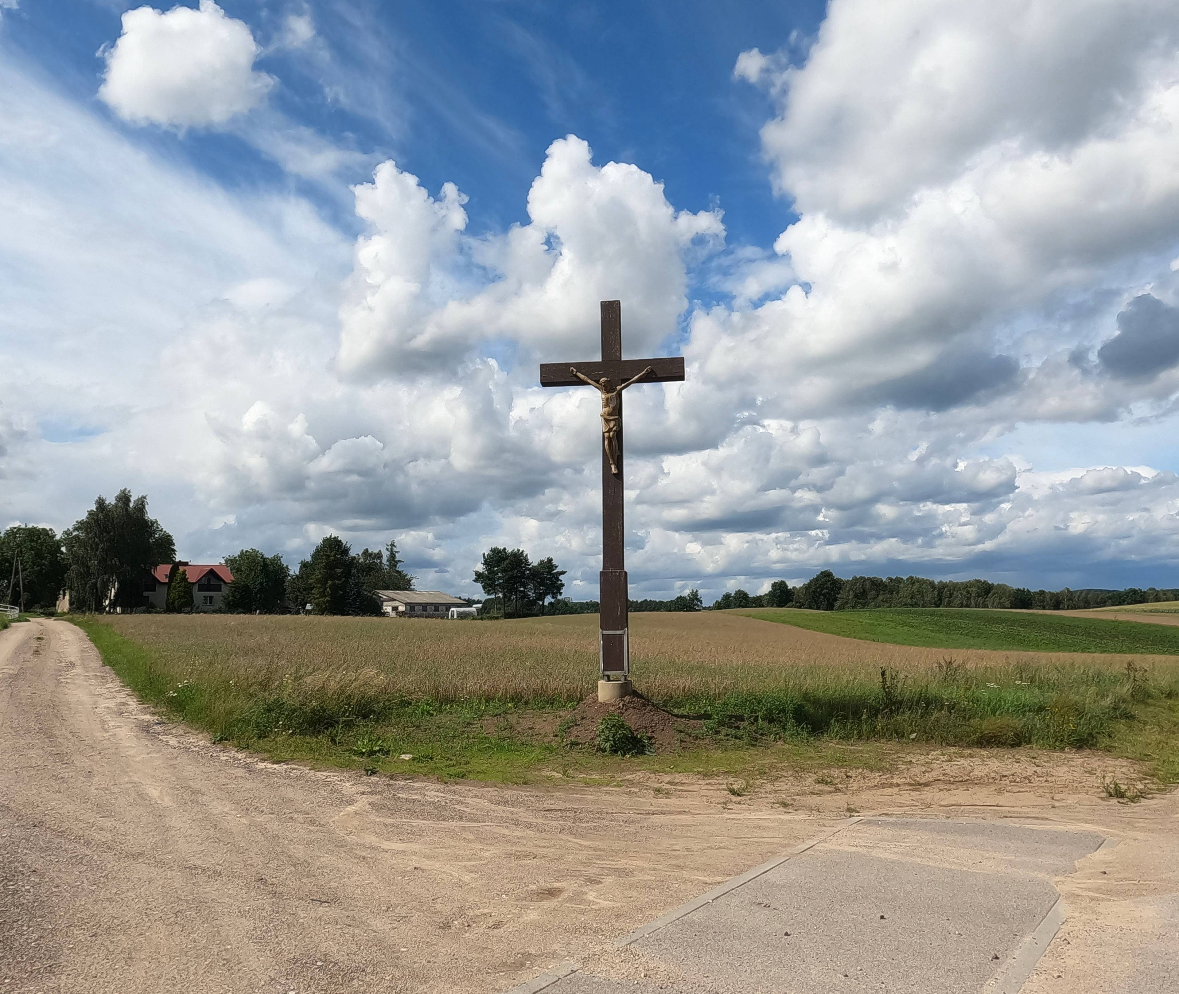 drewniany krzyż na rozstaju dróg, w oddali zabudowania