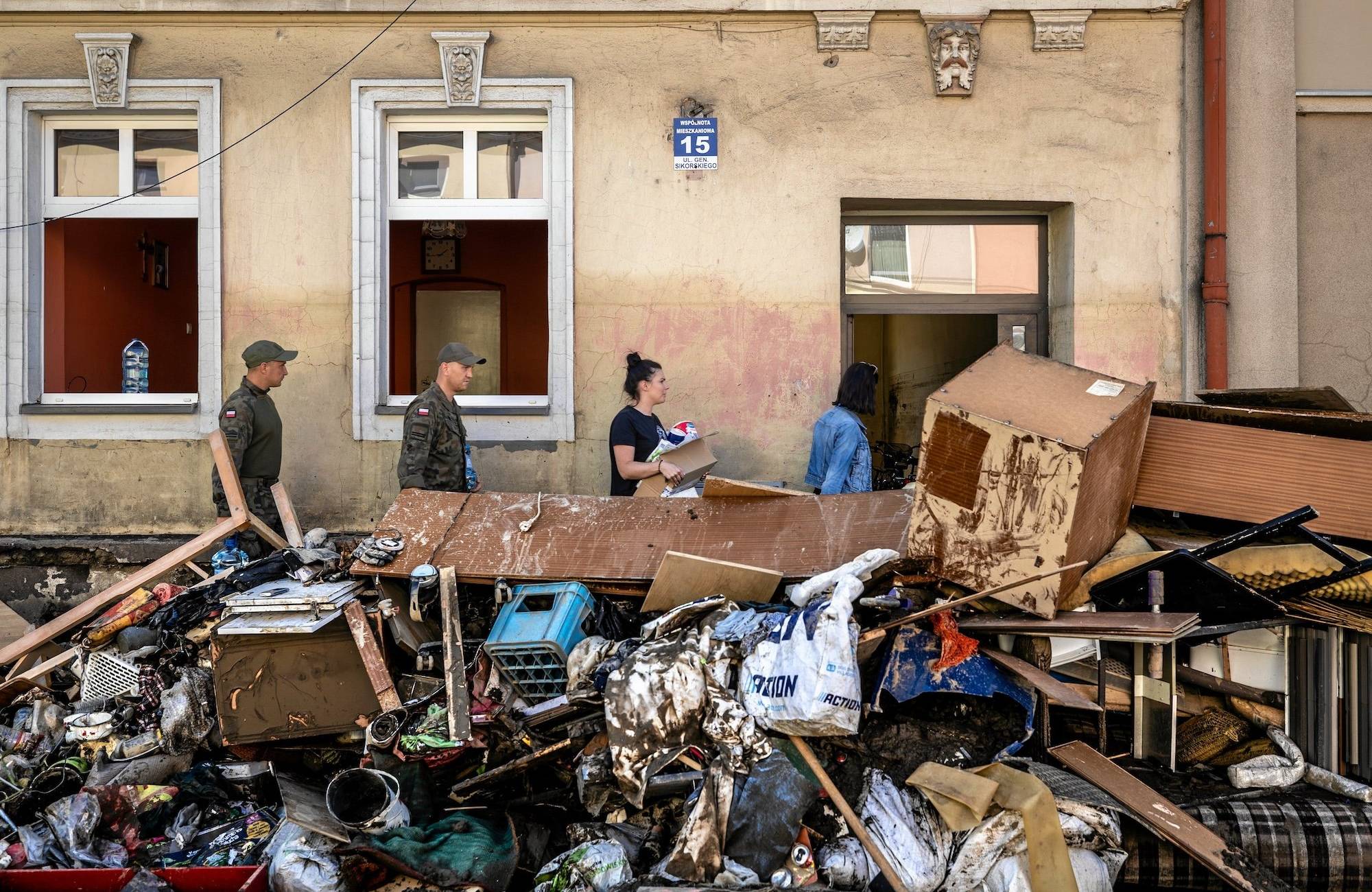 Kilka osób idzie wzdłuż zniszczonego przez powódź budynku. Na pierwszym planie przedmioty zniszczone przez wodę