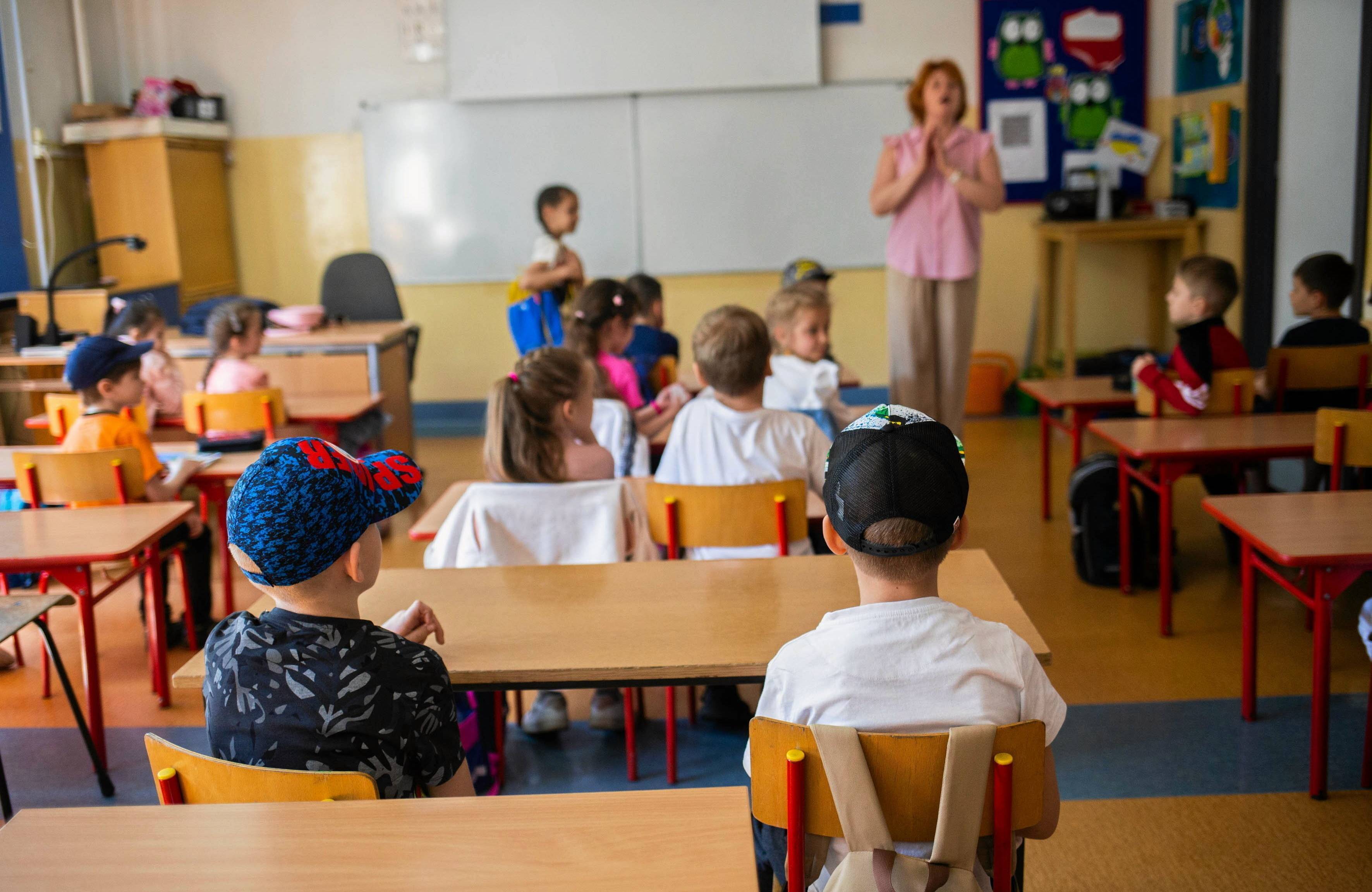 Na zdjęciu są dzieci w wieku ok. siedem lat. Siedzą za szkolnymi ławkami. W tle obok tablicy jest nauczycielka. Ma różową bluzkę i rude włosy
