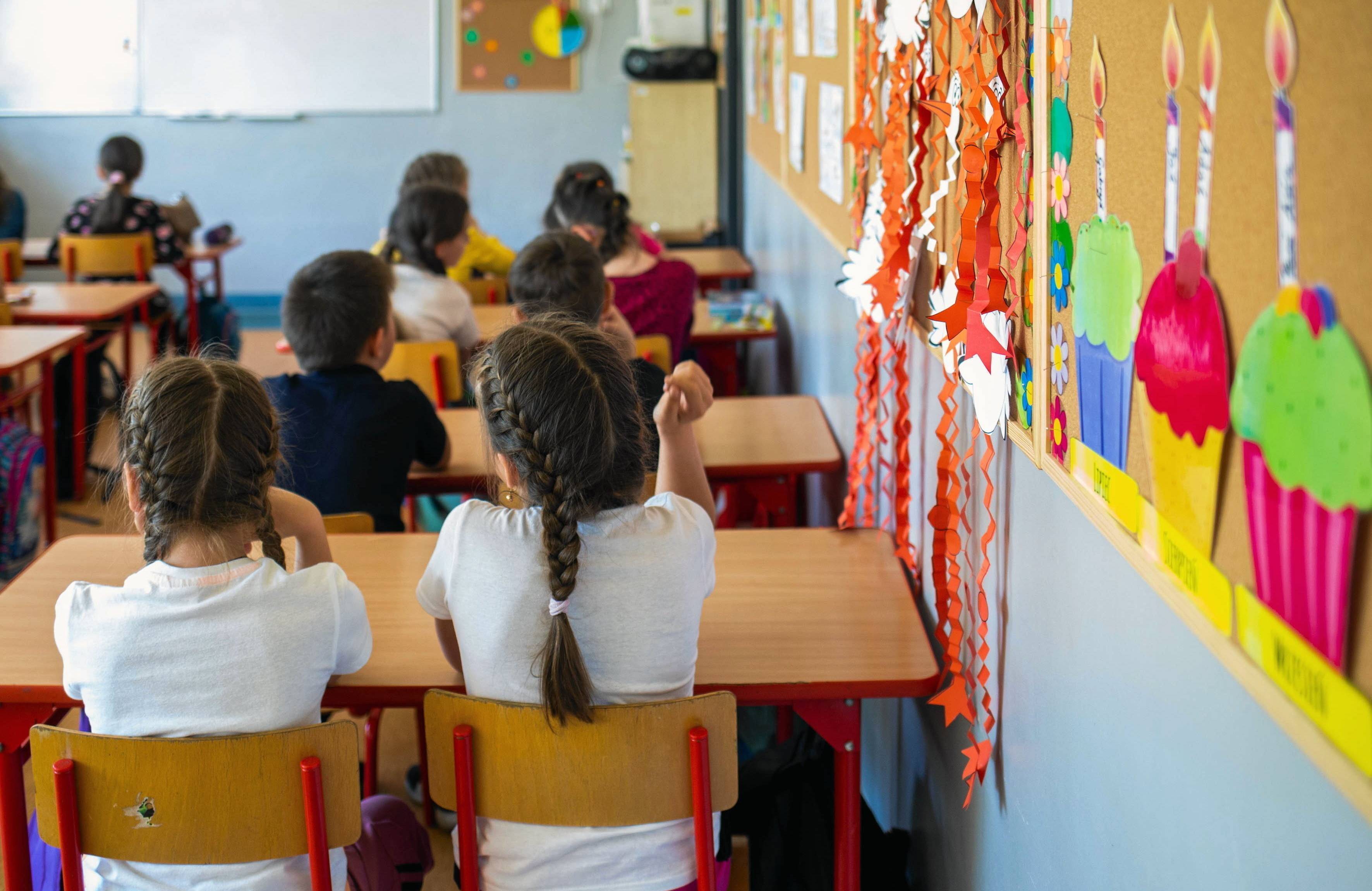 Na zdjęciu są dzieci siedzące za ławkami. Najbliżej widać dziewczynki z brązowymi warkoczykami. Siedzą tyłem do kamery. Są ubrane w białe bluzki.