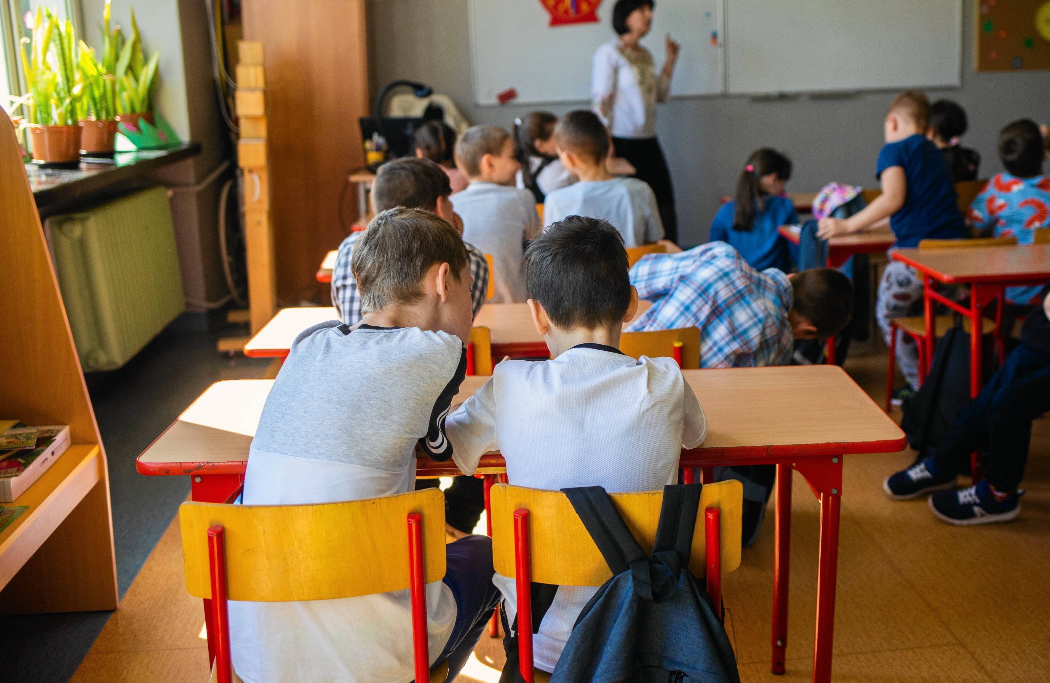 Na zdjęciu są dzieci, które siedzą za ławkami. W tle obok tablicy jest kobieta w białej koszuli. Dzieci siedzą tyłem do kamery
