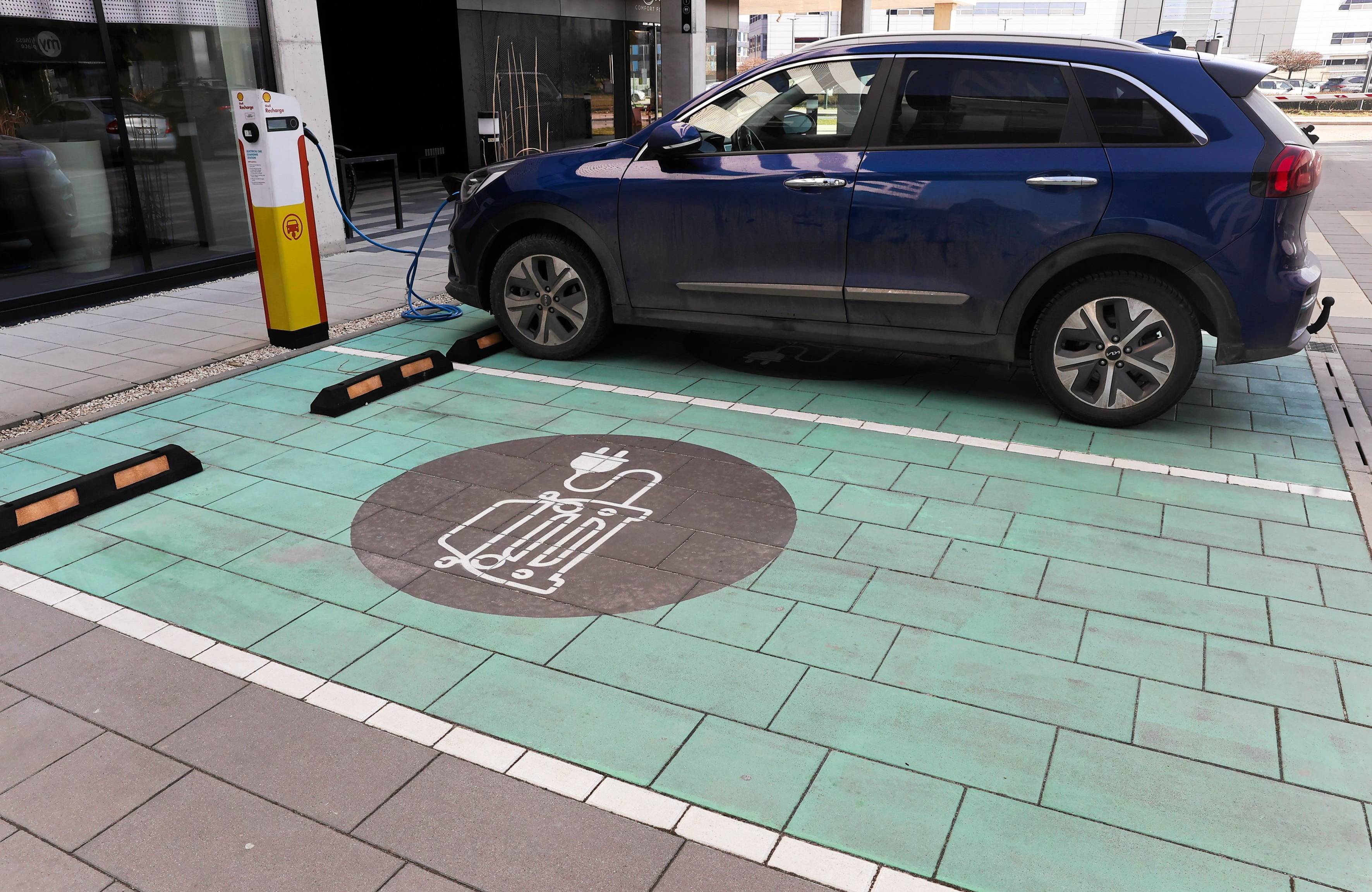 Zielony prostokąt namalowany na płytach chodnikowych z piktogramem samochodu. W tle stacja ładowania pojazdu elektrycznego