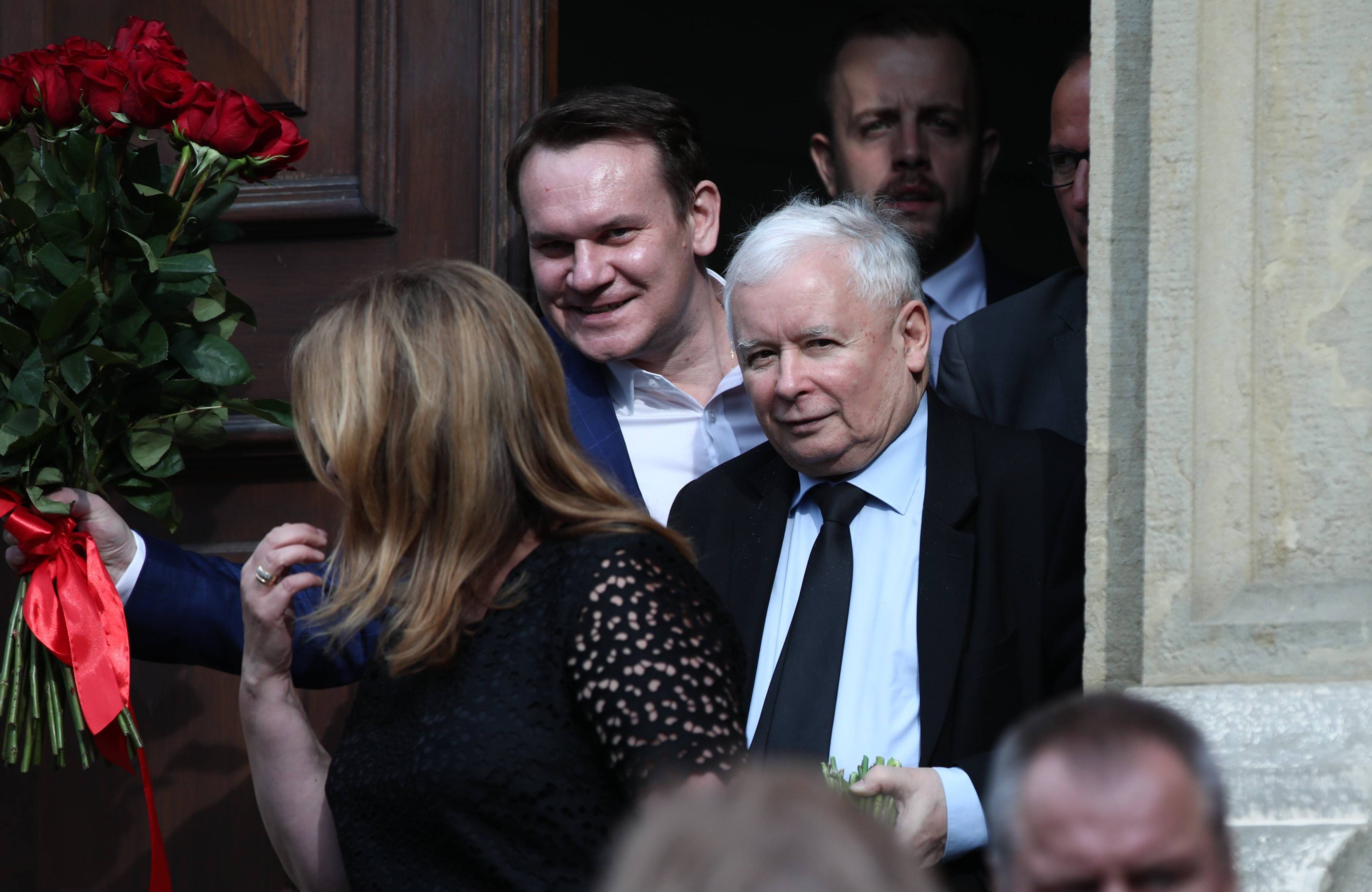 Dwóch uśmiechniętych mężczyzn, młodszy i starszy, wychodzi z budynku w otoczeniu innych osób dookoła