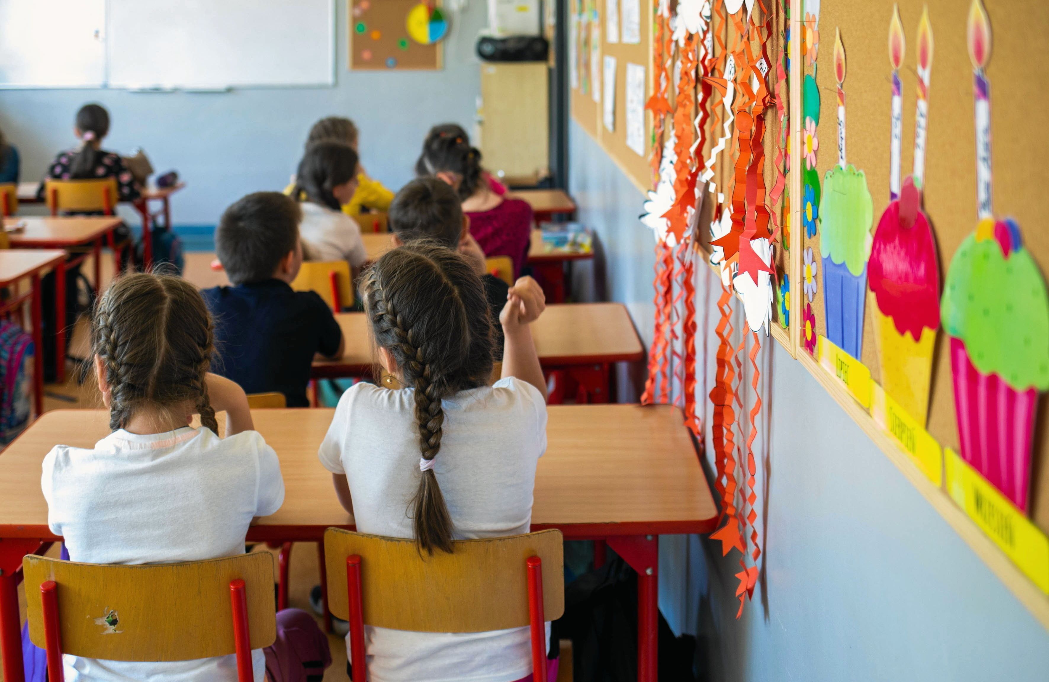 Na zdjęciu są dzieci siedzące za ławkami. Najbliżej widać dziewczynki z brązowymi warkoczykami. Siedzą tyłem do kamery. Są ubrane w białe bluzki.