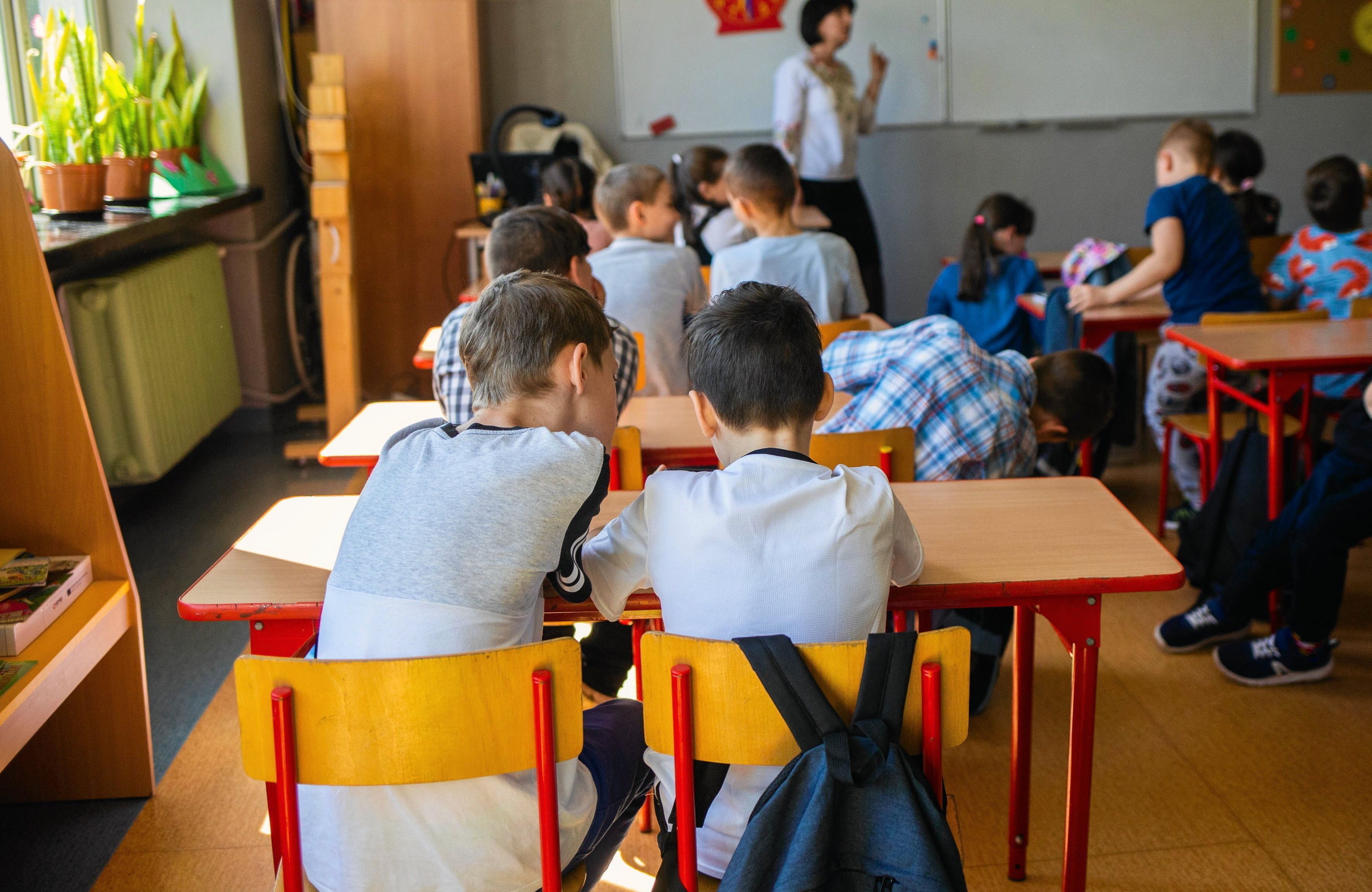 Na zdjęciu są dzieci, które siedzą za ławkami. W tle obok tablicy jest kobieta w białej koszuli. Dzieci siedzą tyłem do kamery