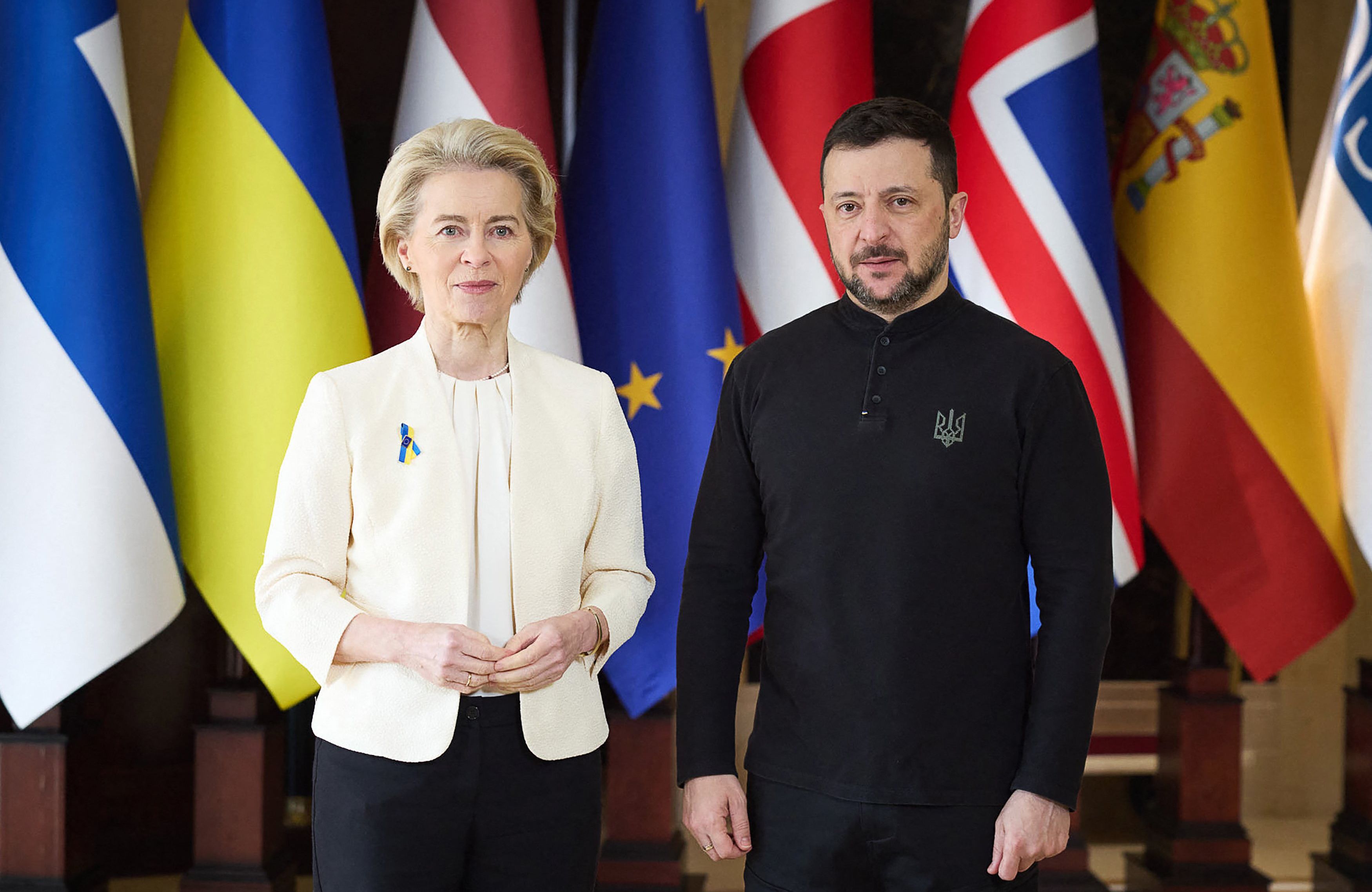 Kobieta w kremowej marynarce (Przewodnicząca KE Ursula von den Leyen) i brodaty mężczyzna w bluzie z symbolem trójzębu na piersi (prezydent Ukrainy Wołodymyr Zełenski) pozują na tle europejskich flag