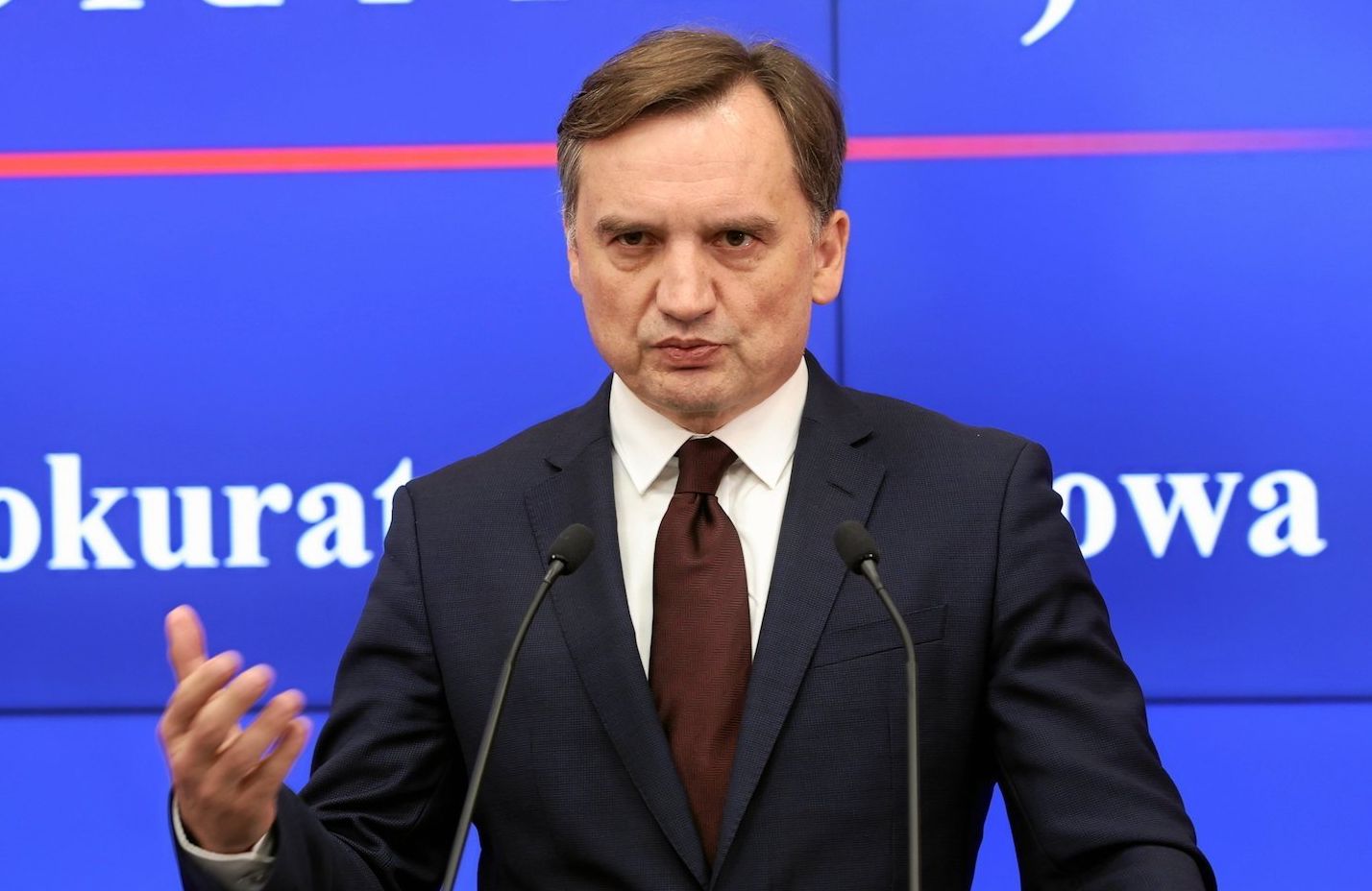 Prokurator Generalny i Minister Sprawiedliwości Zbigniew Ziobro na konferencji w Prokuraturze Krajowej. Przemawia z pulpitu na tle banerów PK ubrany w ciemny garnitur i bordowy krawat.