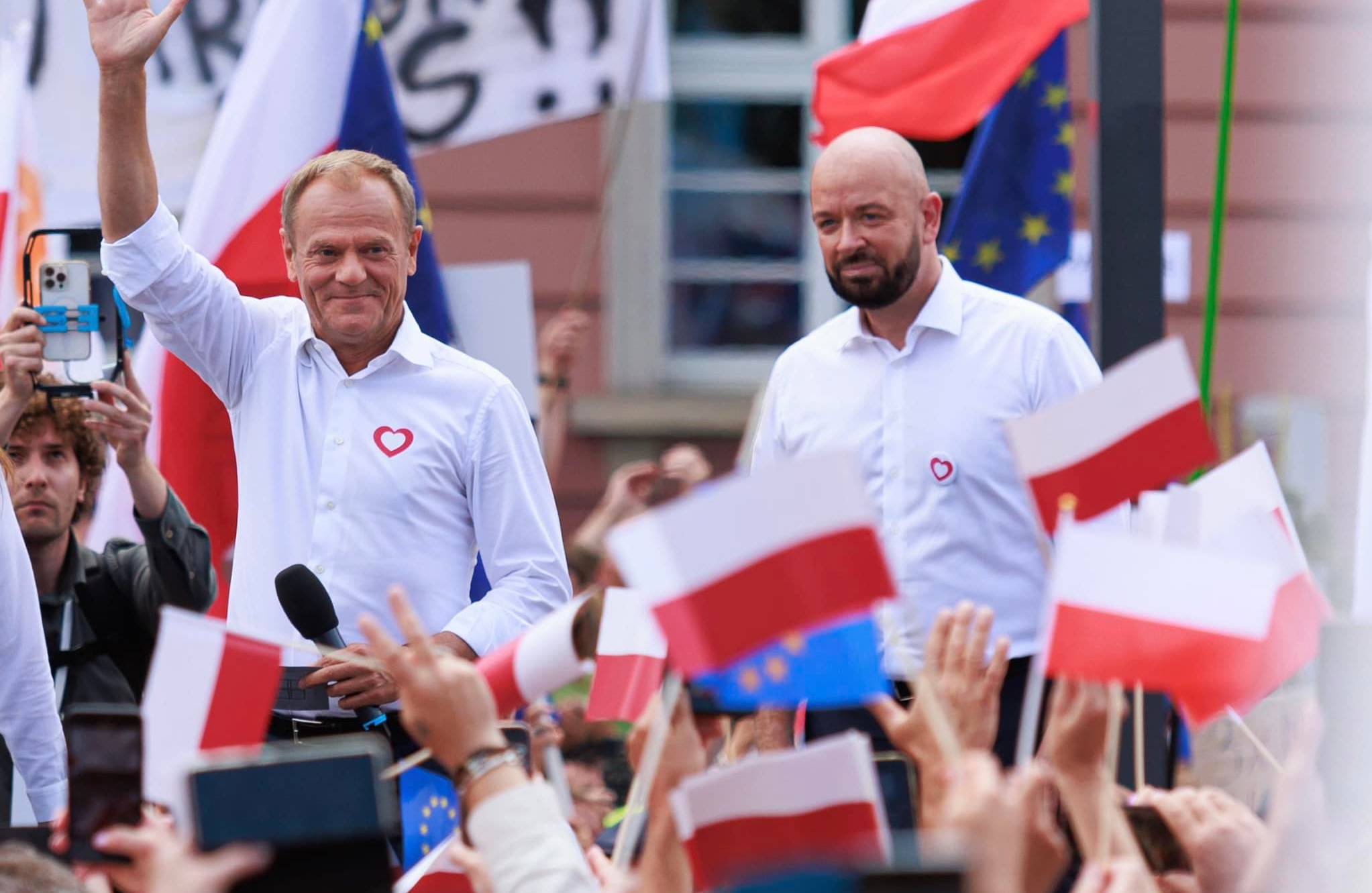 Dwóch mężczyzn w białych koszulach z małymi naklejkami czerwonych serduszek na piersi macha i uśmiecha się do tłumu wyborców machających biało-czerwonymi flagami