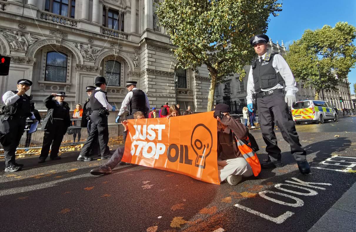 aktywiści z banerem just stop oil siedzą na ulicy otoczeni przez policję