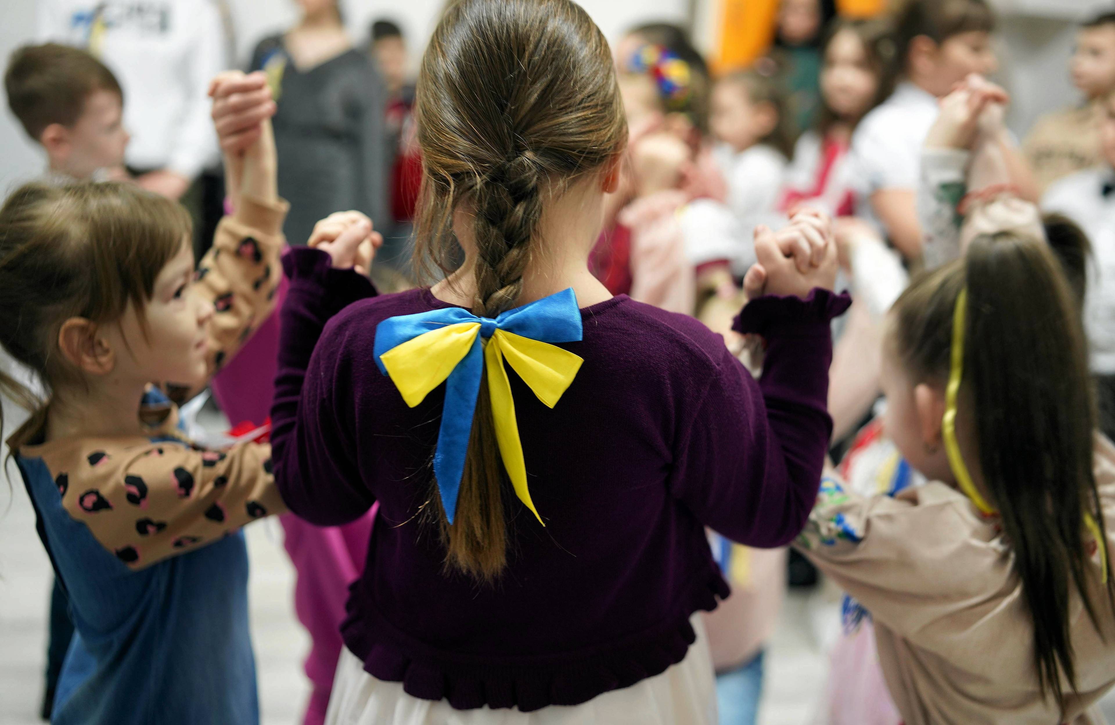 Na zdjęciu są dzieci w wieku 6-7 lat. Po środku tyłem stoi dziewczynka. Jest ubrana w bordową bluzkę, na wakoczyku ma zawiązaną kokardę w kolorach niebiesko-żółtych. Ma brązowe włosy